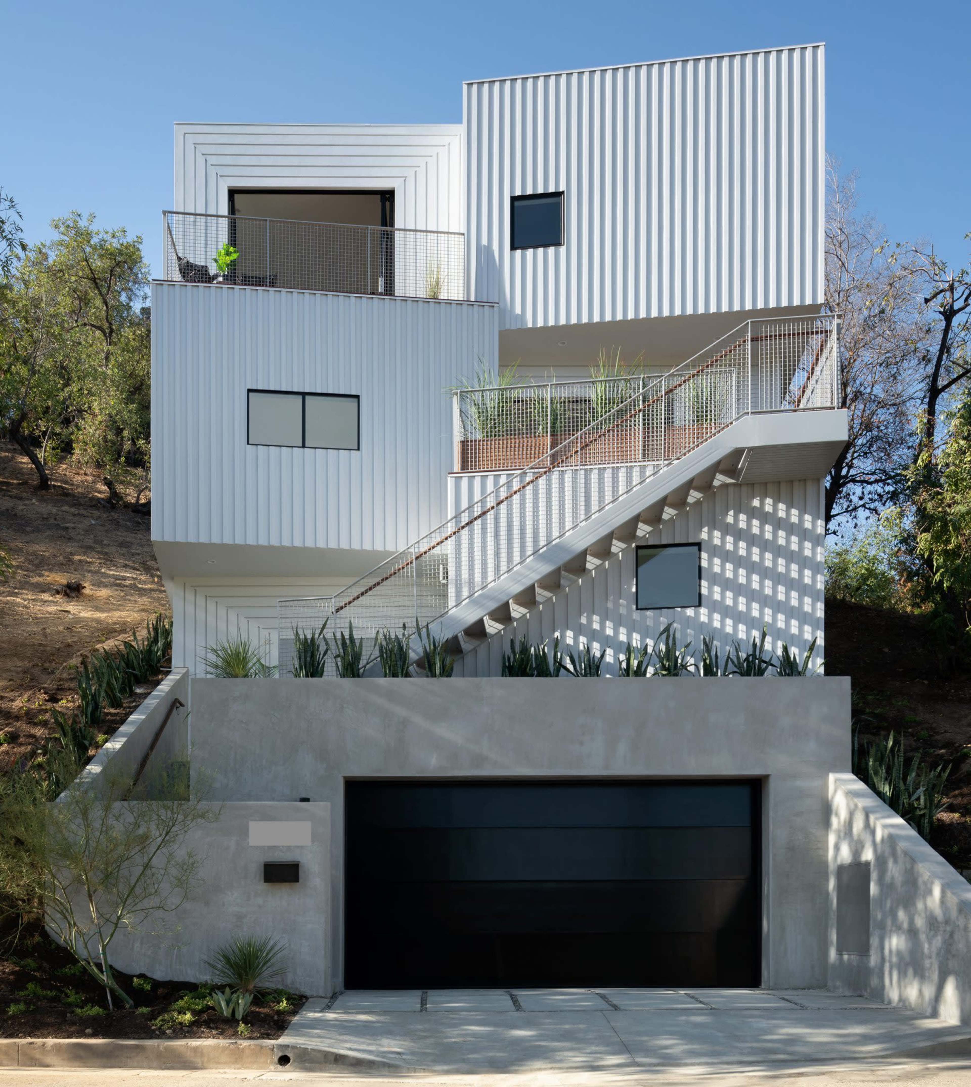 A modern three-story house with a white corrugated exterior, featuring multiple balconies and a landscaped entrance.
