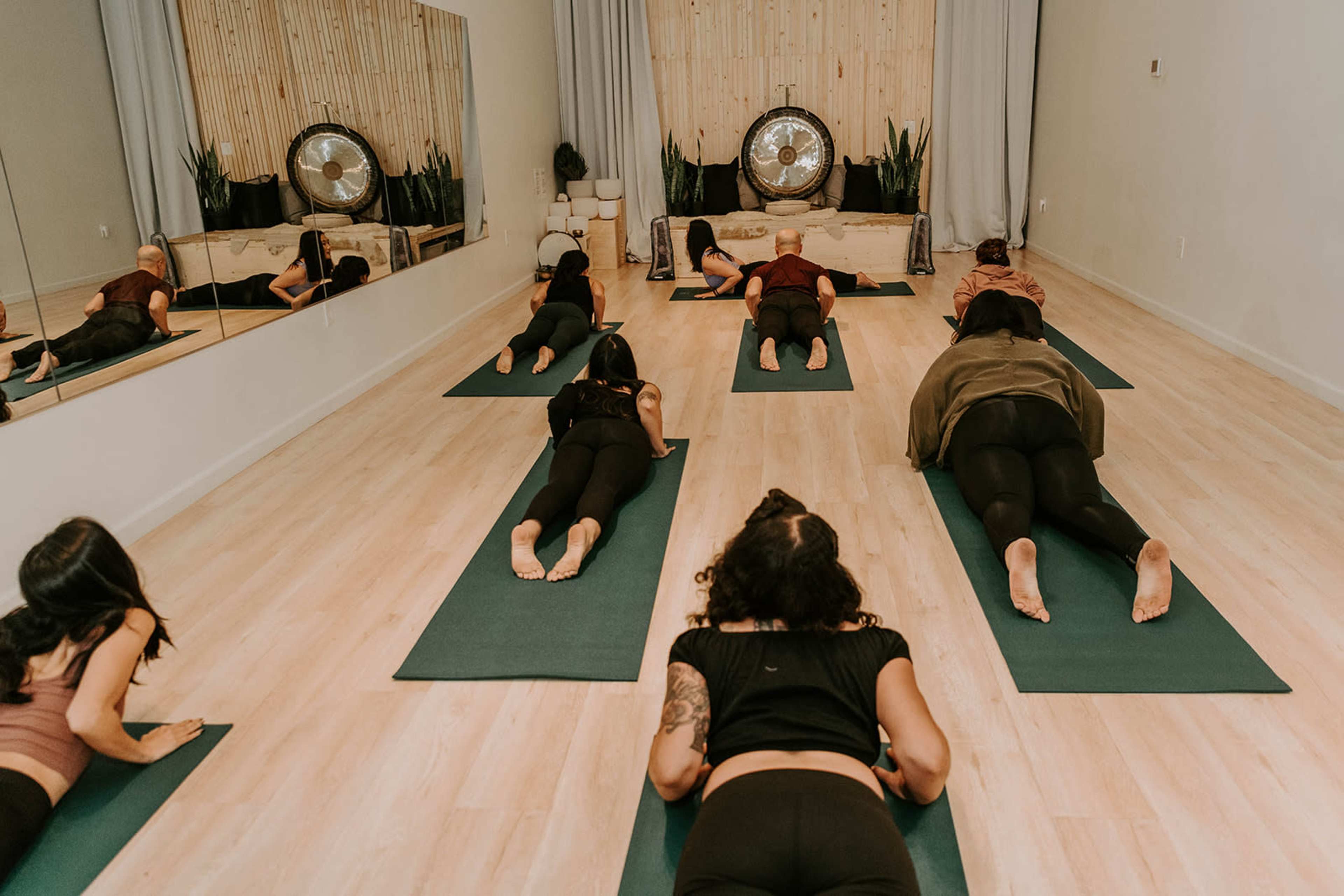 A group of people practices yoga on mats in a studio with a wooden wall and a large gong in the background.