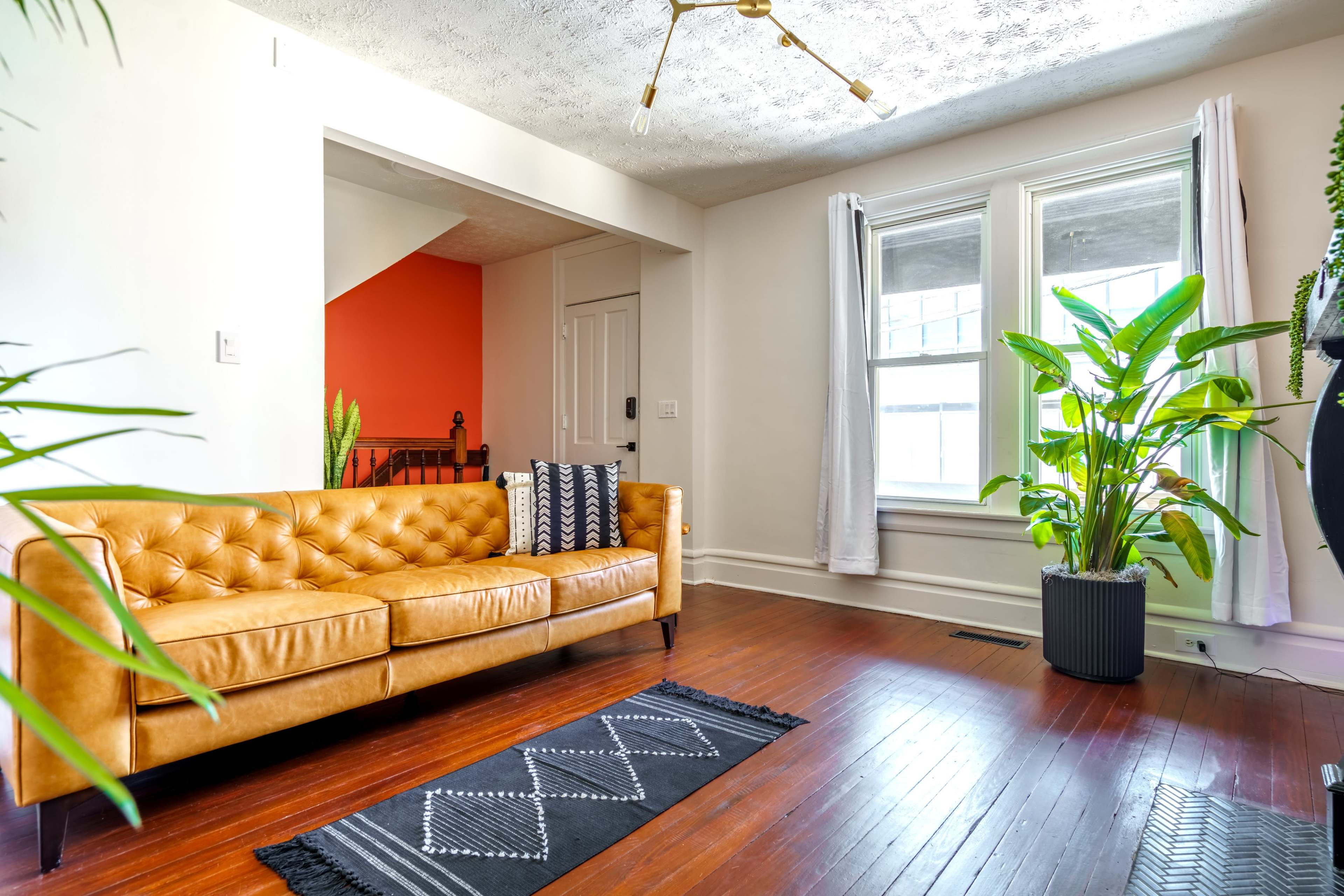 The image shows a living room featuring a brown leather sofa, a black patterned rug, and large windows with natural light illuminating the space.