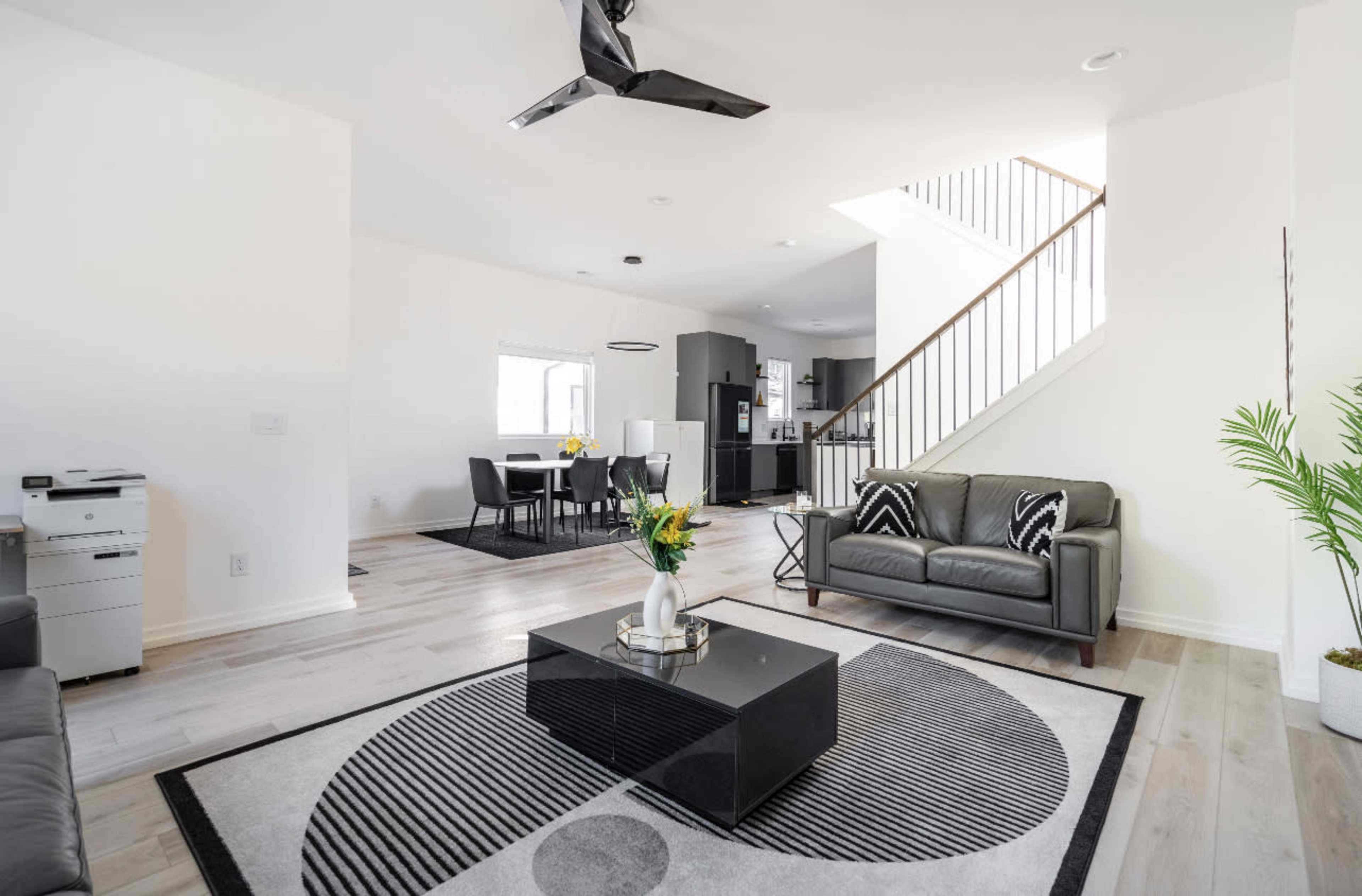 A modern living room with a black coffee table, two leather sofas, and a staircase leading to an upper level, alongside a dining area and kitchen visible in the background.