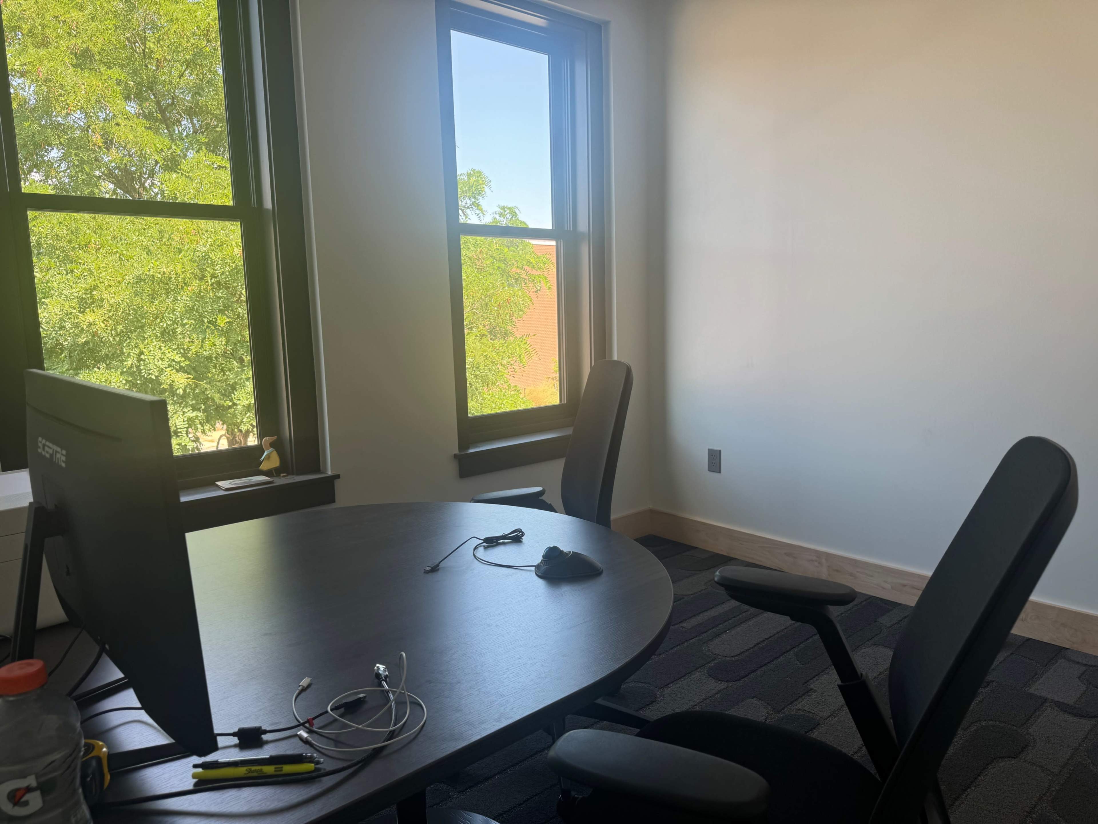 The image shows a small office space with a round table, a computer monitor, a mouse, and two black chairs positioned by the windows.