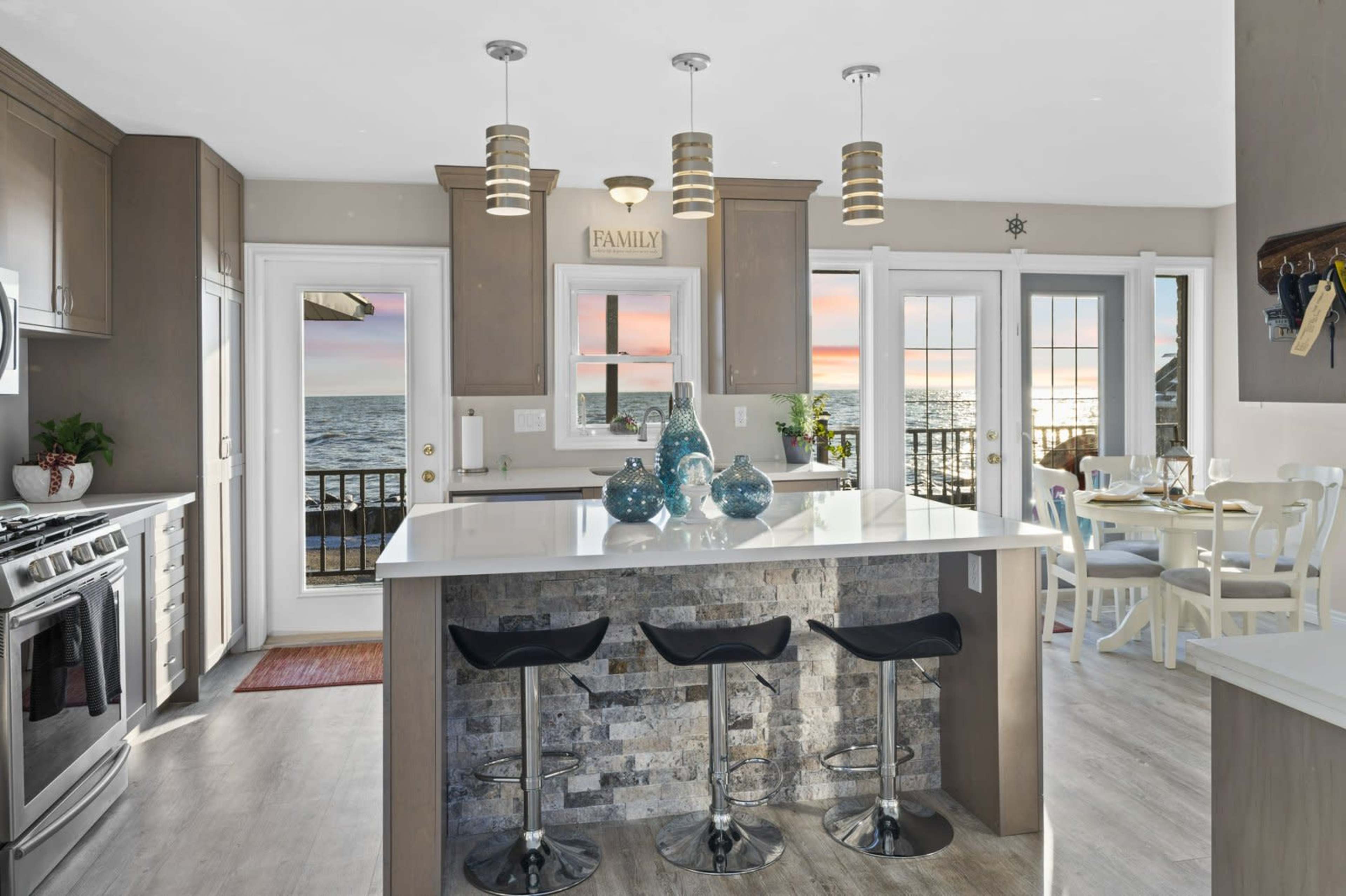 The kitchen features a central island with three bar stools, modern appliances, and large windows that offer a view of the ocean and a sunset.