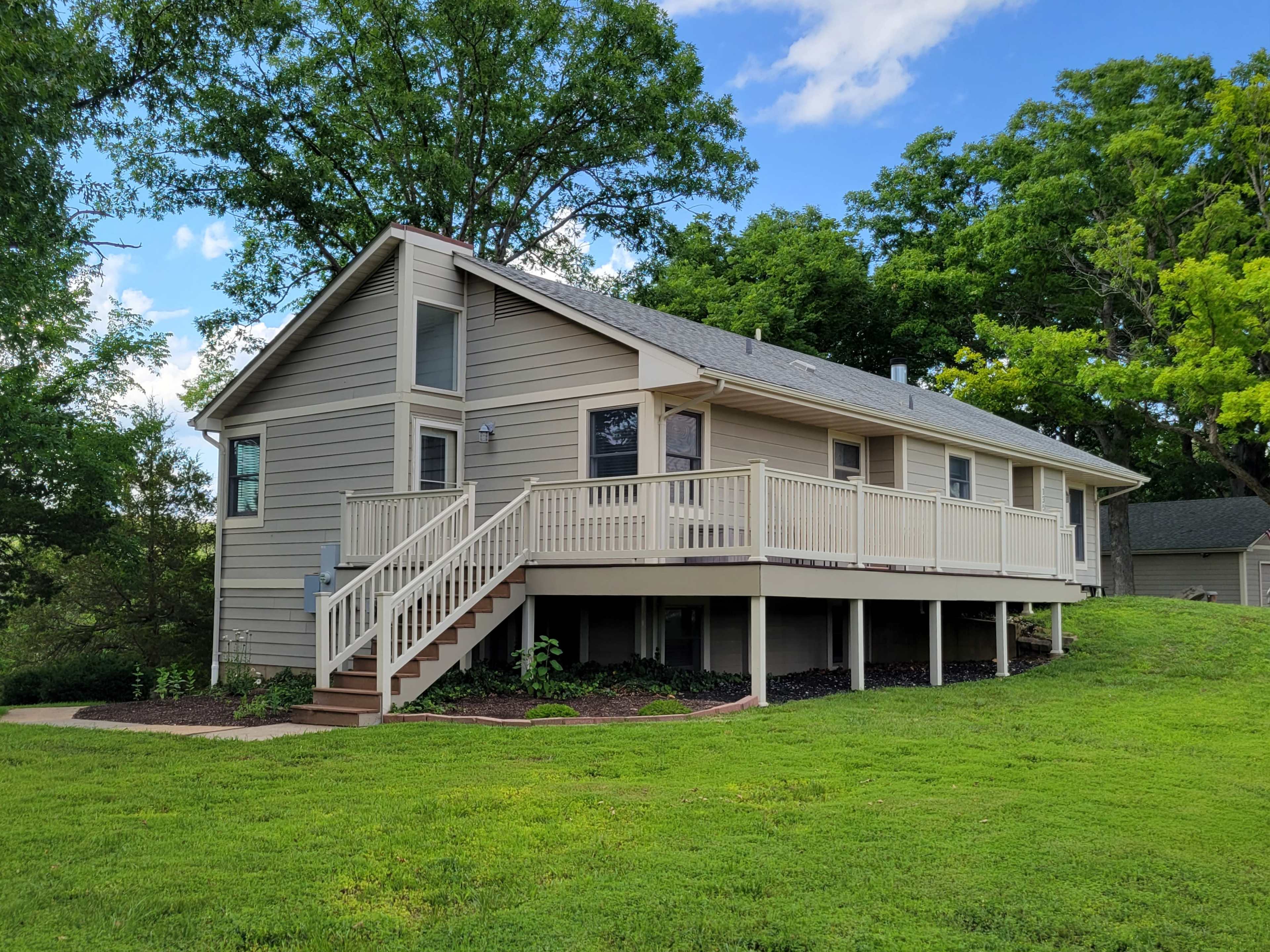 A single-story house with a light-colored exterior and a wrap-around deck sits on a grassy slope surrounded by trees.