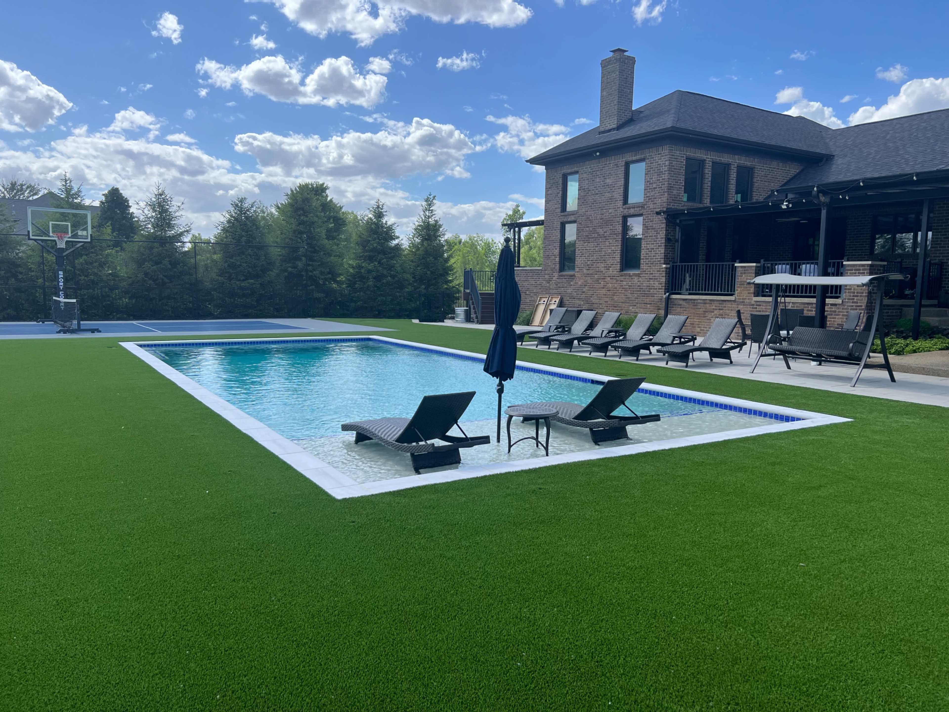 A backyard with a paved swimming pool surrounded by lawn chairs, a basketball court in the background, and a two-story brick house.
