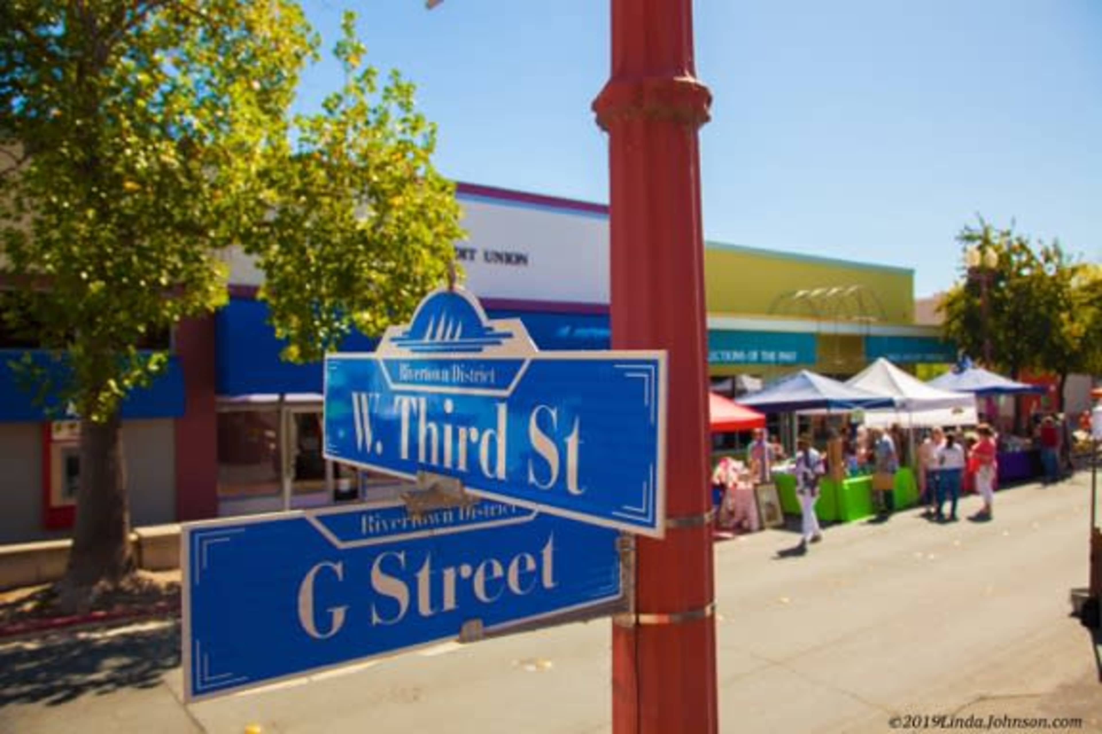 A street sign at the intersection of W. Third St. and G Street is visible, with colorful market tents set up nearby under clear blue skies.