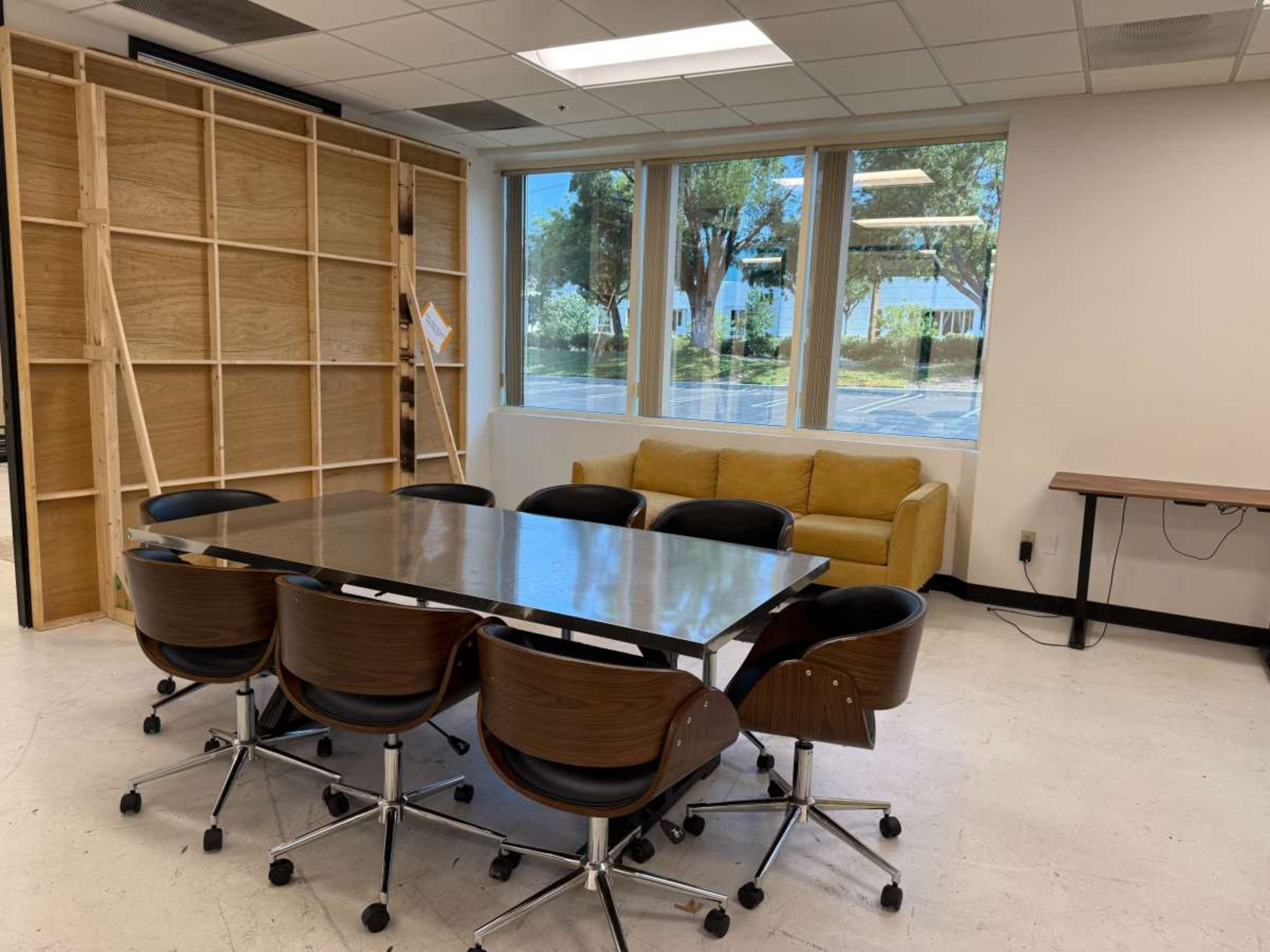 The image shows a meeting room with a large table surrounded by black chairs, a yellow couch in the background, and large windows letting in natural light.