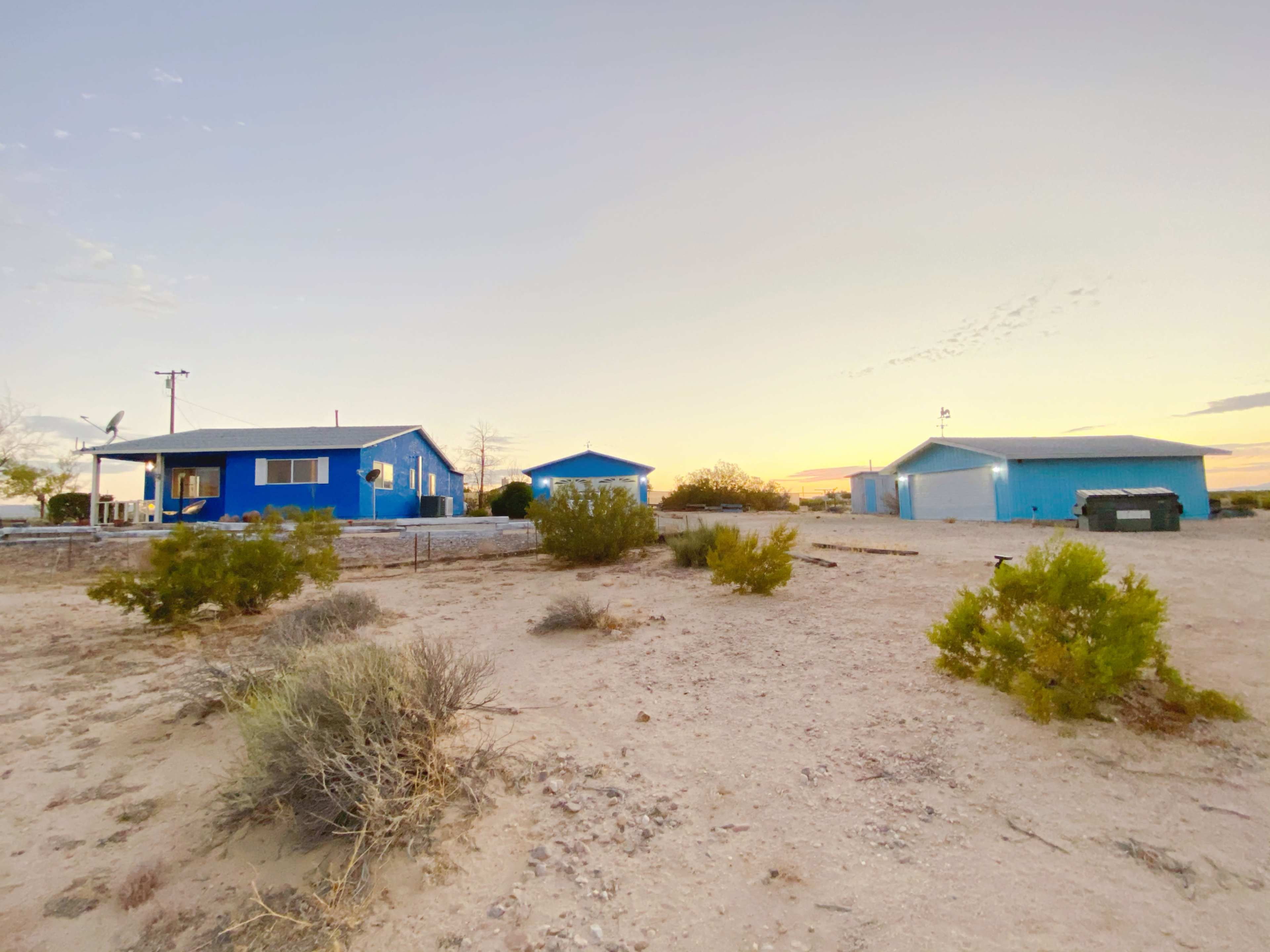A blue house and an adjacent building are set against a desert landscape at sunset, with sparse vegetation around them.