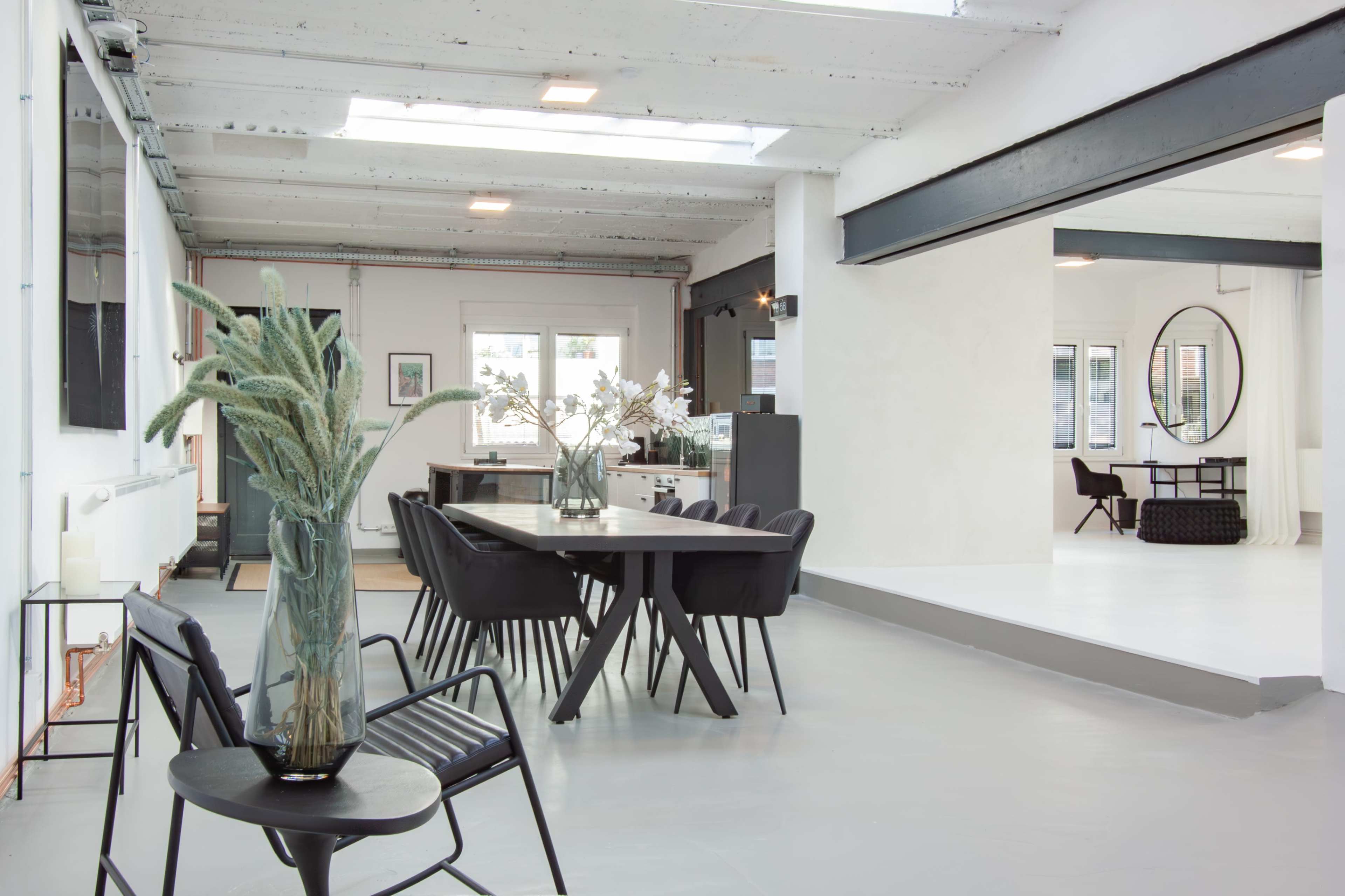 A spacious, modern interior features a dining table surrounded by black chairs, with a large window providing natural light and a minimalist design aesthetic.