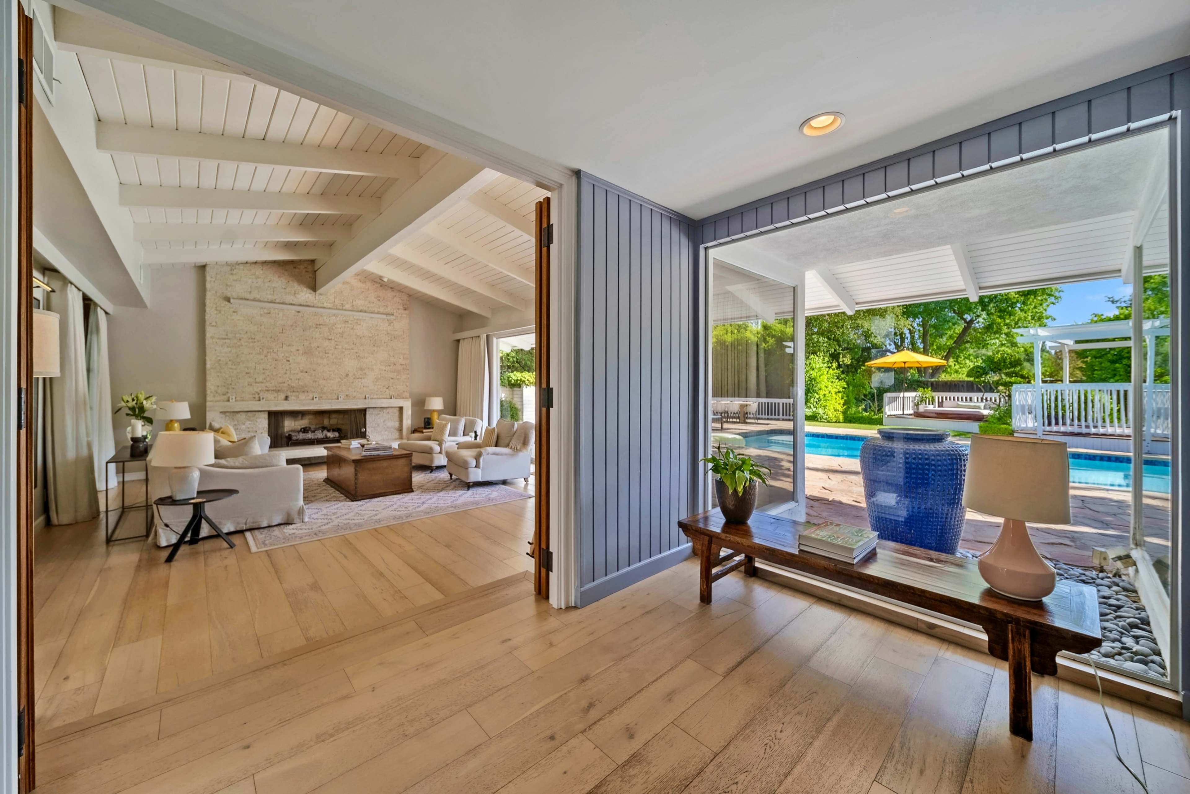 A spacious living area features a stone fireplace, large windows, and a view of a poolside patio.