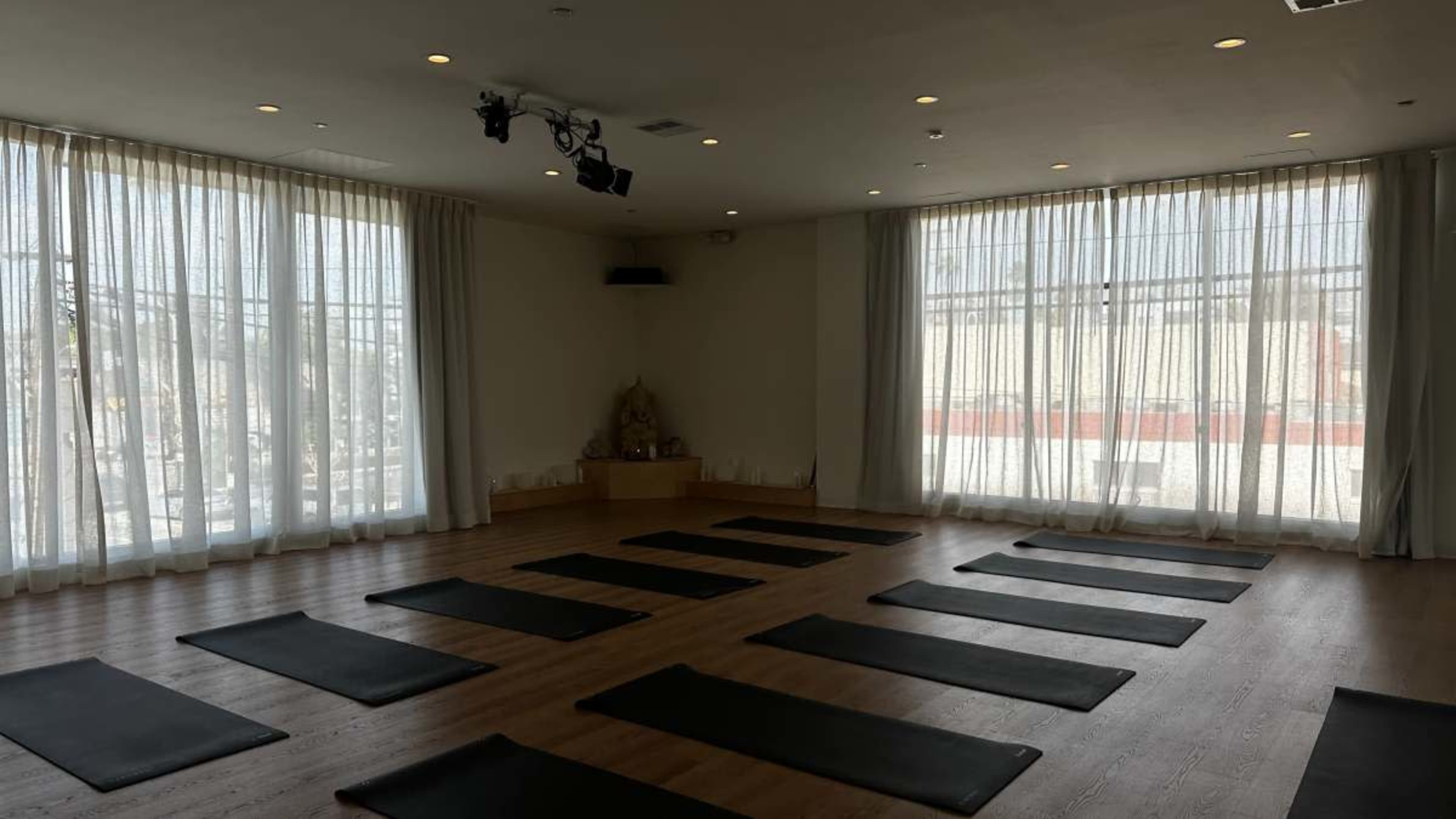 The image shows a yoga studio with several mats arranged on a wooden floor, illuminated by natural light coming through large sheer curtains.