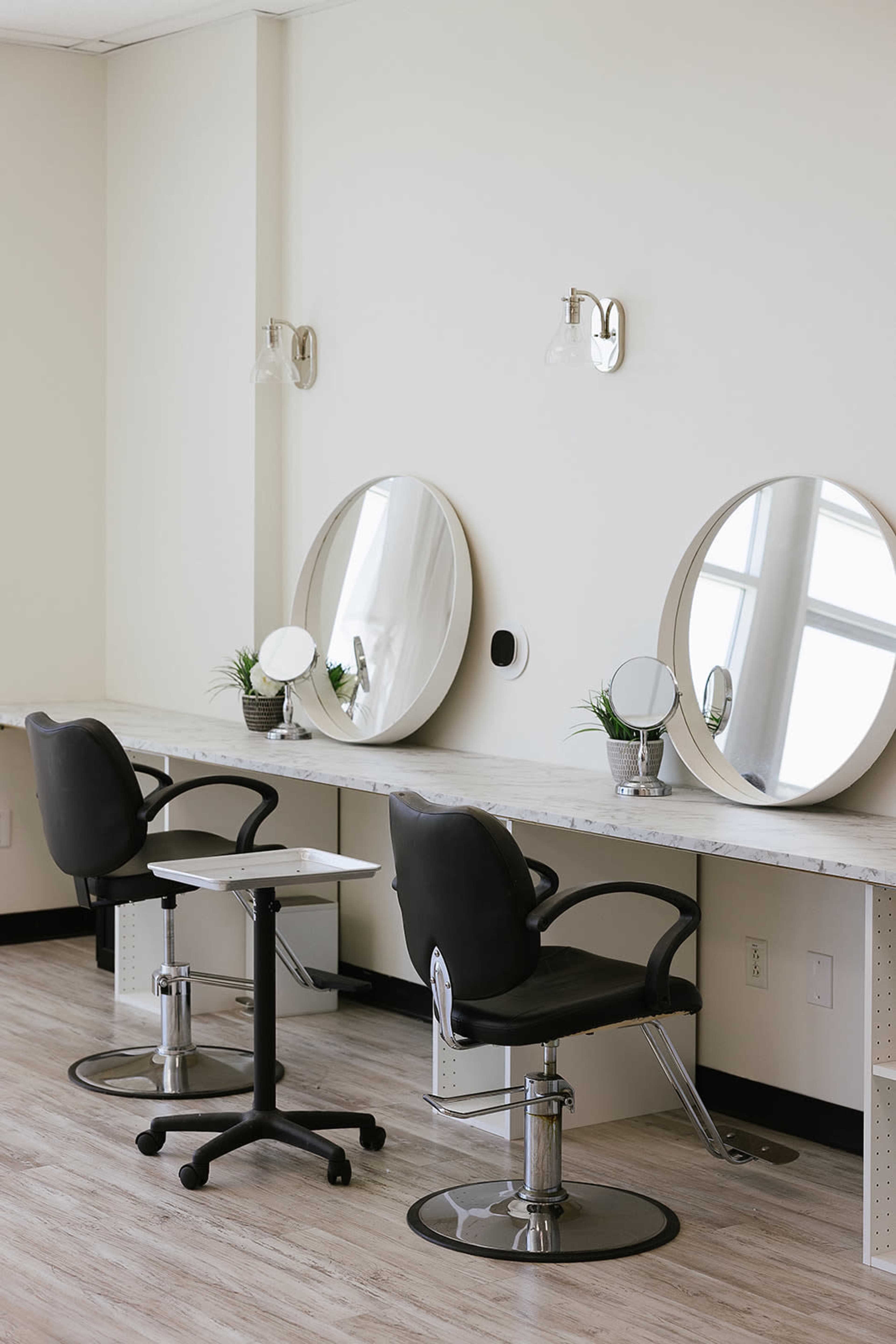 The image shows a minimalist hair salon with two black swivel chairs, round mirrors mounted on the wall, and a small white table in between them.