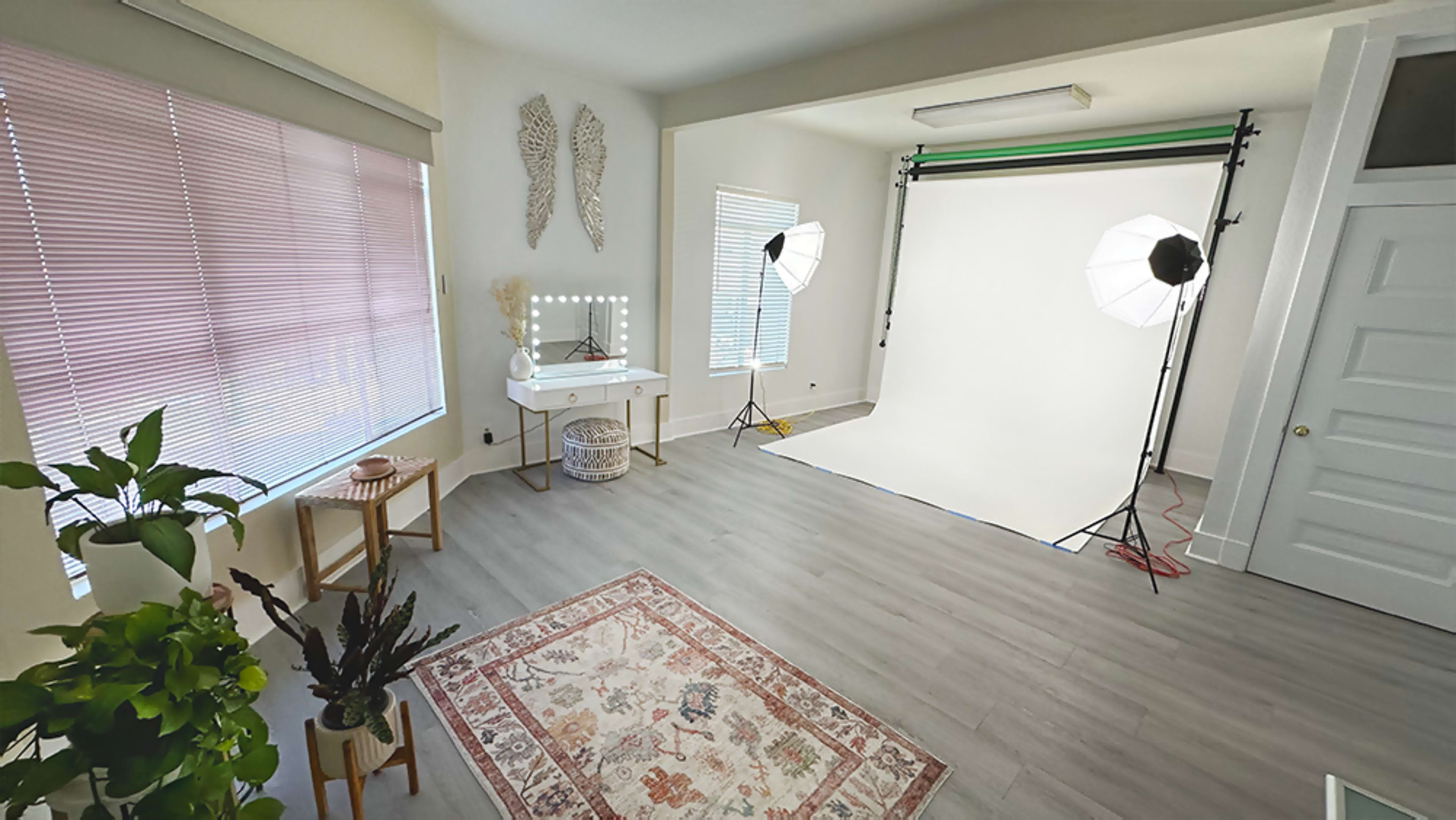 A well-lit studio space with a white backdrop, professional lighting equipment, a vanity table, a small plant, and a decorative rug on the floor.