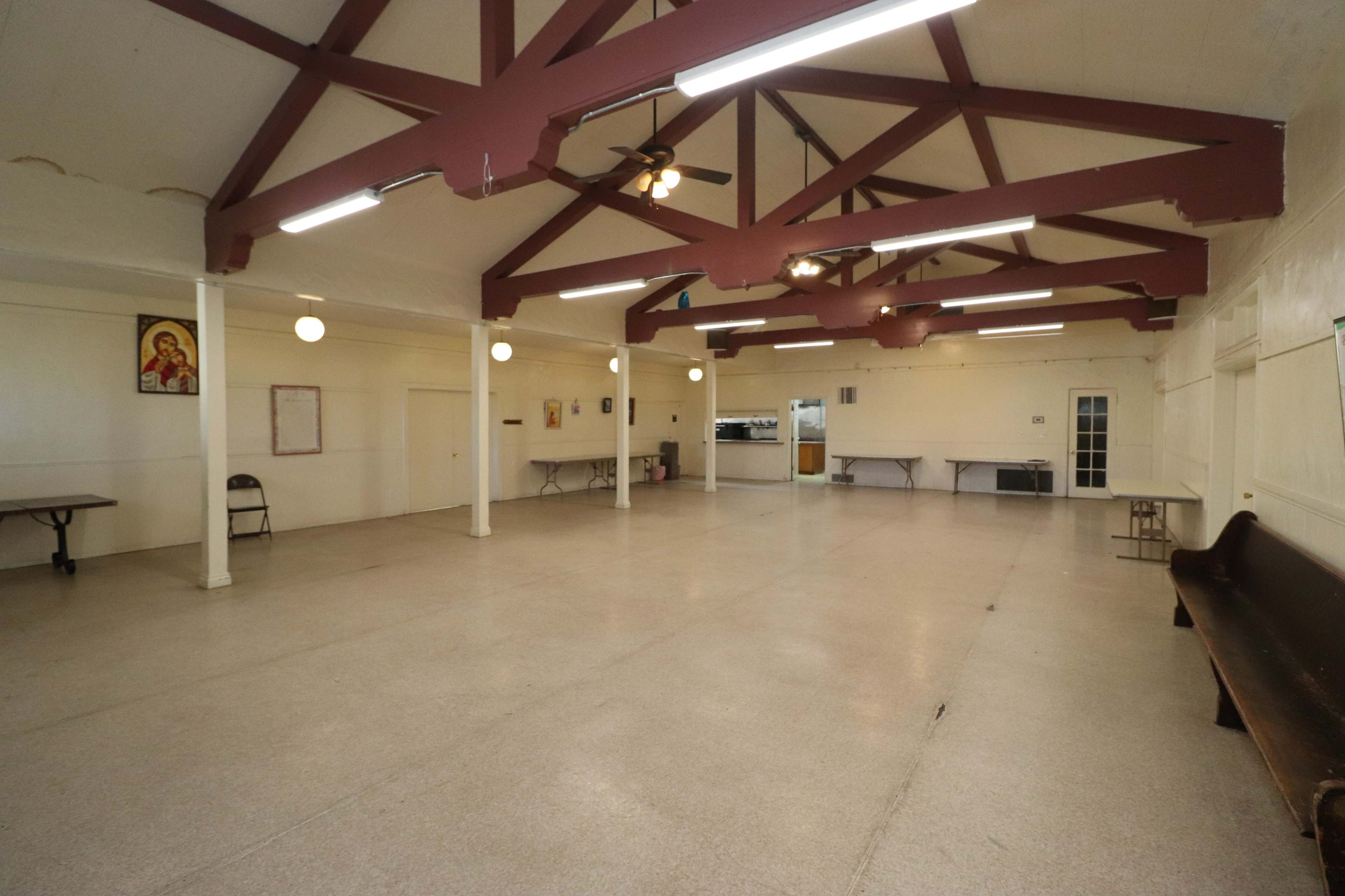 An empty community hall with a high ceiling, exposed beams, and several folding tables along the walls.