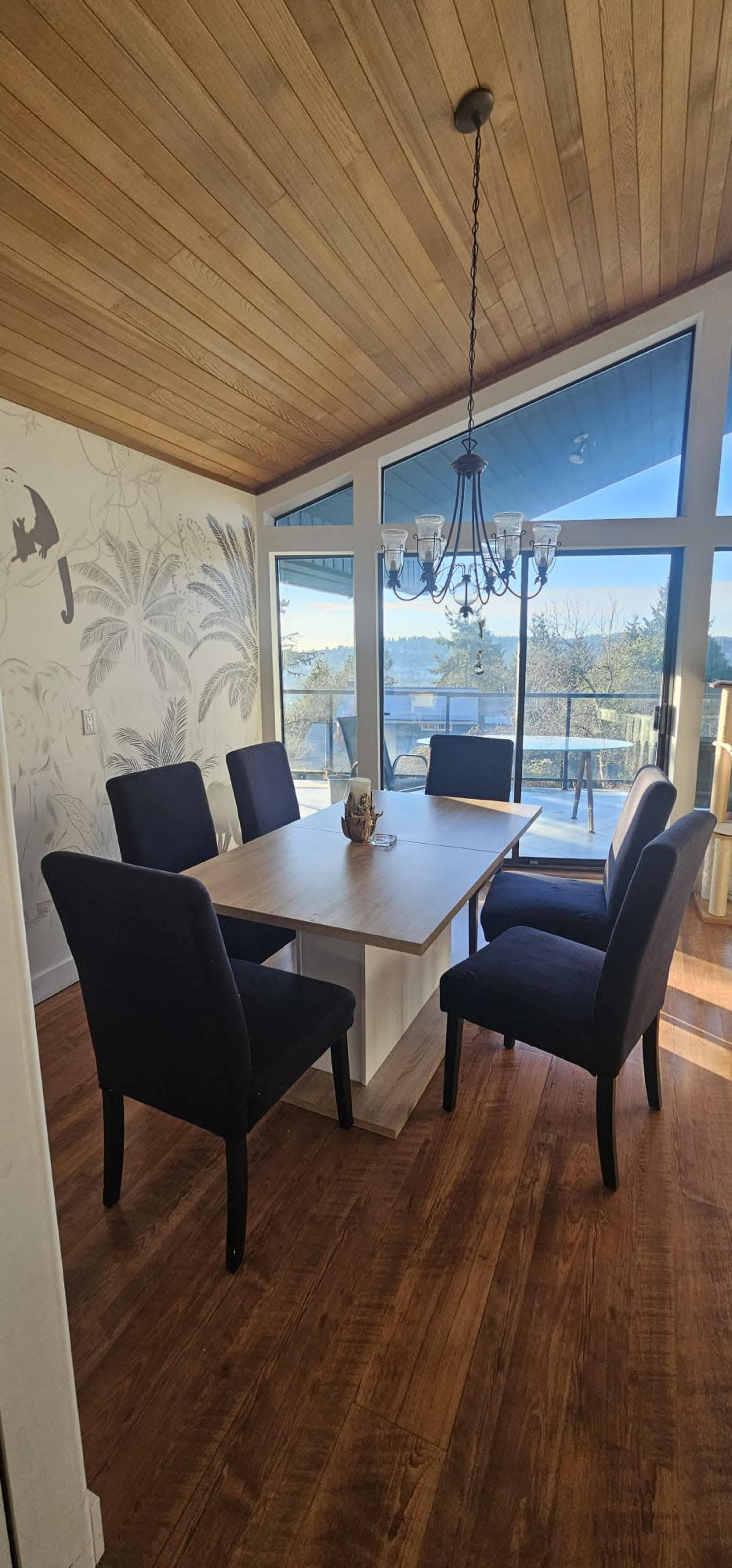 A modern dining area features a wooden table surrounded by four upholstered chairs, with large windows offering a view of outdoor greenery.
