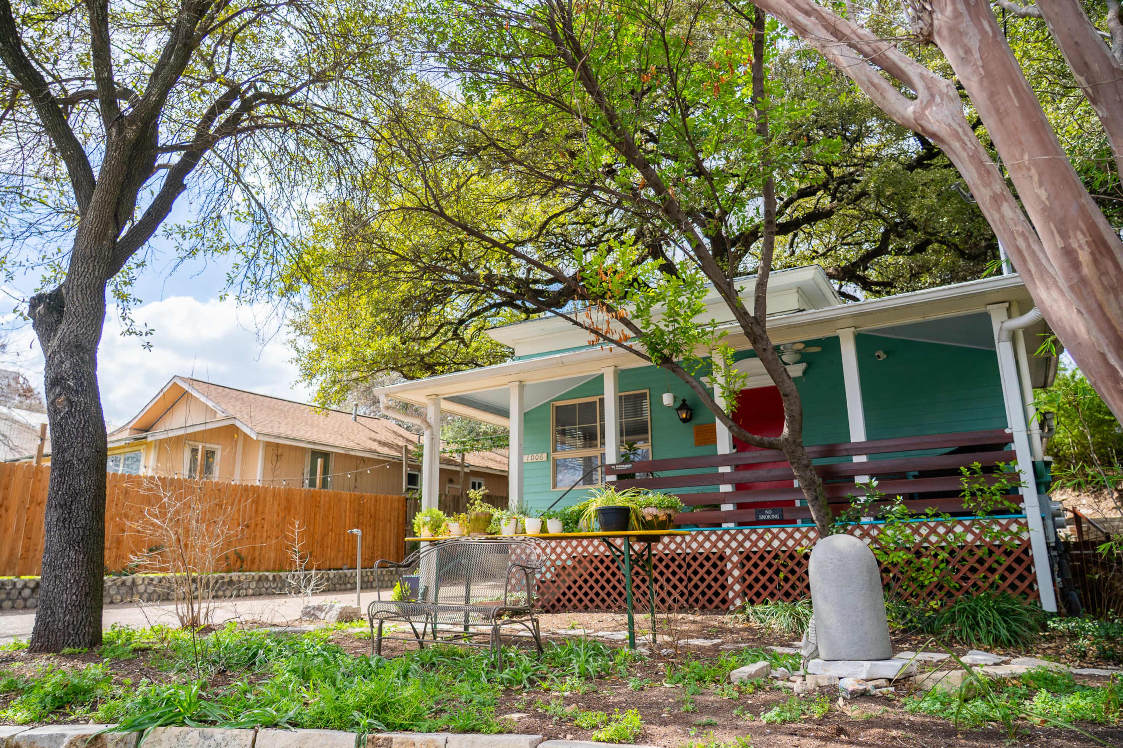 A teal-colored house with a red door sits under lush green trees, accompanied by a small seating area in the front yard.