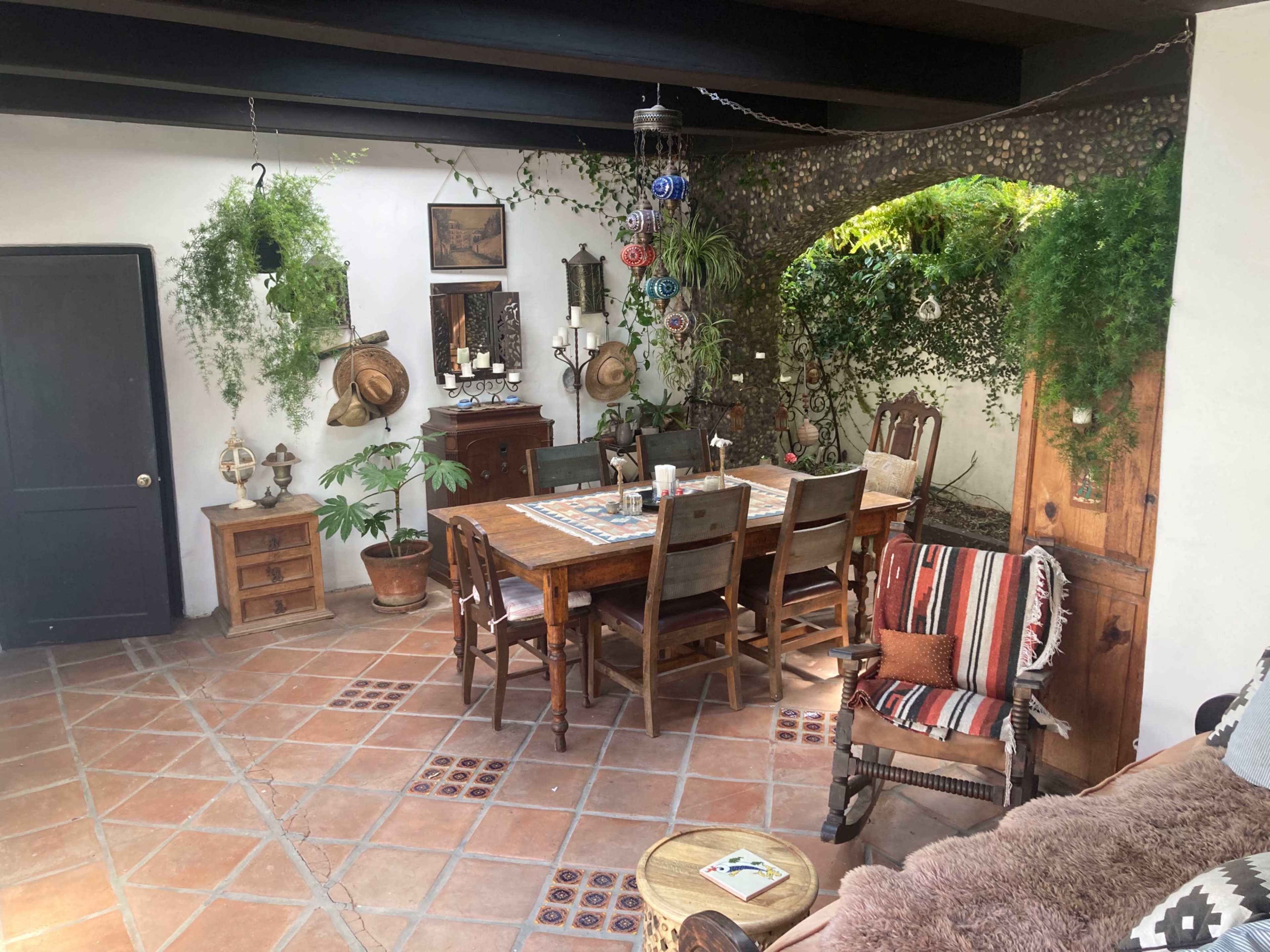 A cozy dining area with a wooden table surrounded by chairs, decorative plants hanging from the ceiling, and various pieces of rustic furniture.