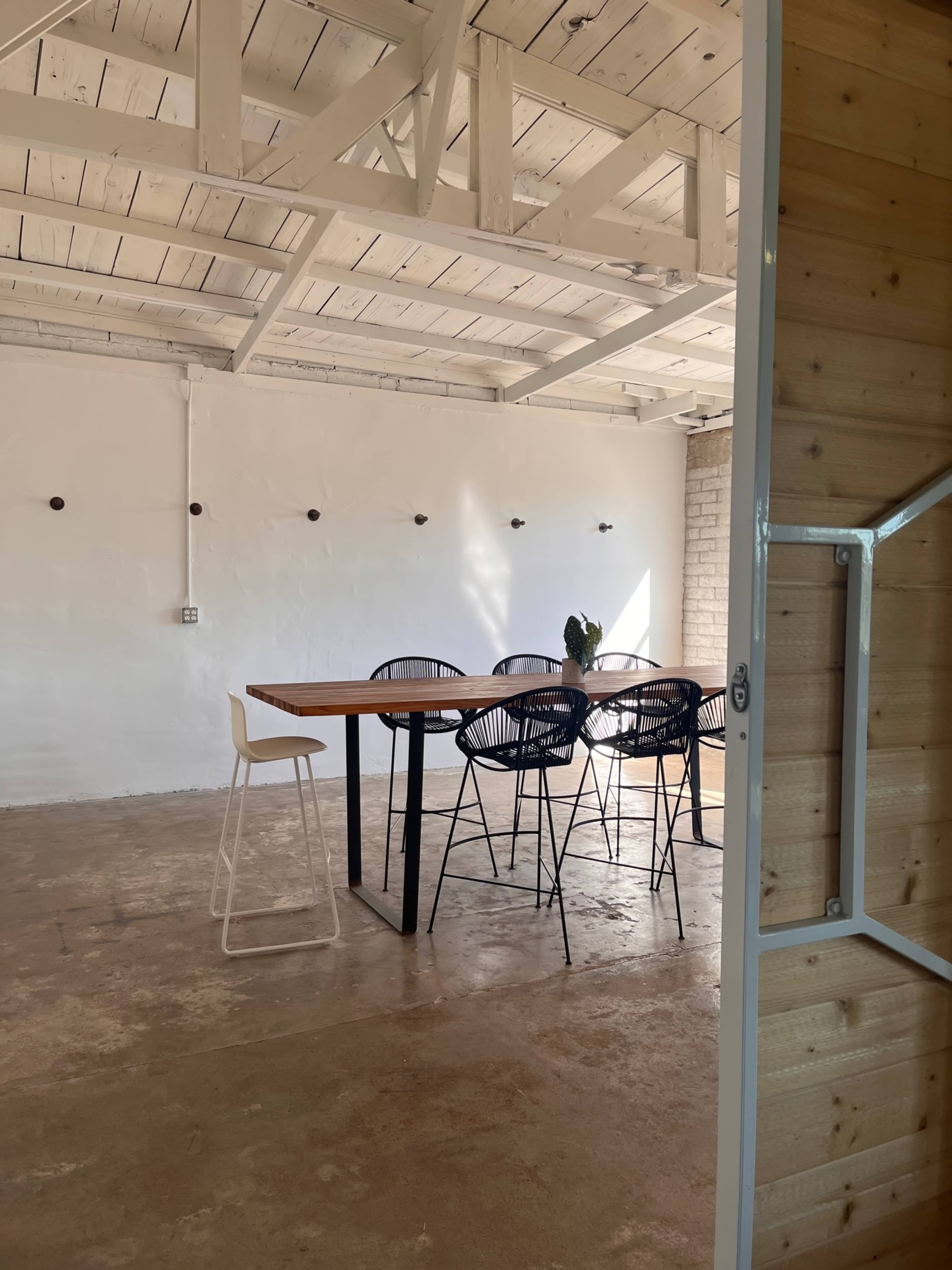 A minimalistic meeting room features a wooden table surrounded by black wire chairs, with natural light illuminating the space through exposed beams and a white wall.
