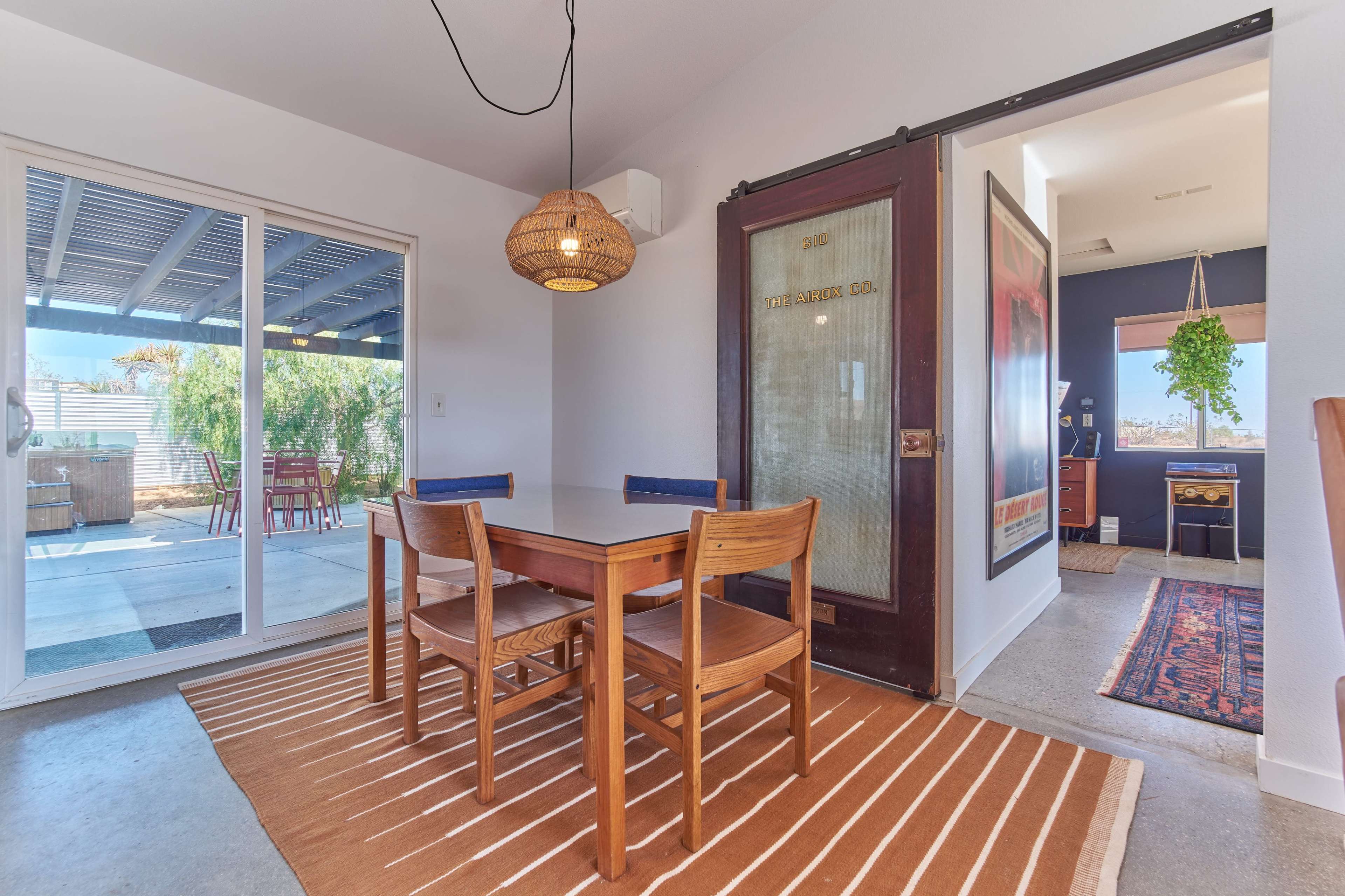 A dining area features a wooden table with four chairs, a woven light fixture, and a sliding glass door that leads to an outdoor space.