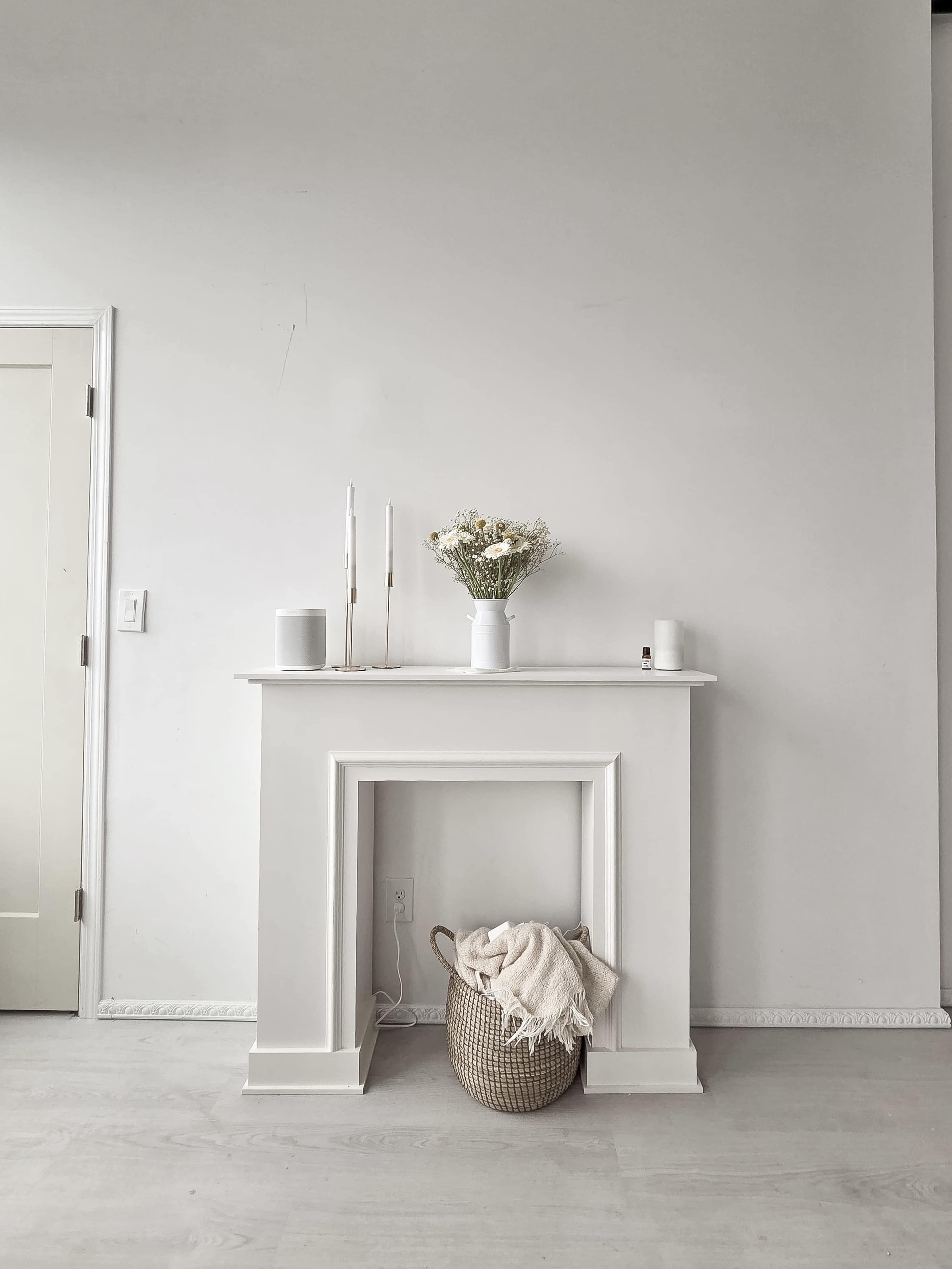 A white mantel sits against a blank wall, with a vase of flowers on top and a woven basket beneath it.