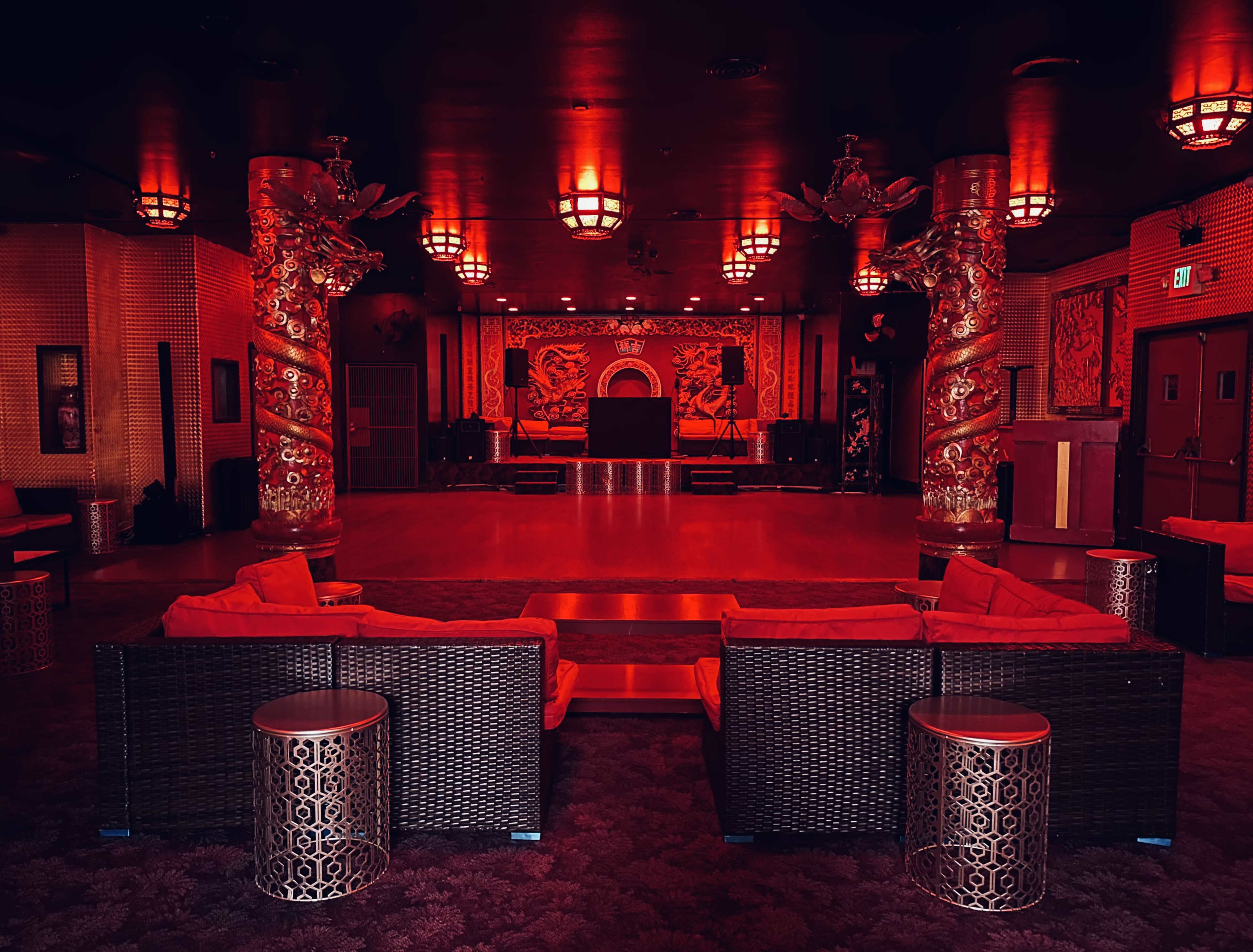 The image depicts an empty lounge area with red lighting, featuring decorative pillars, plush seating, and a stage in the background.