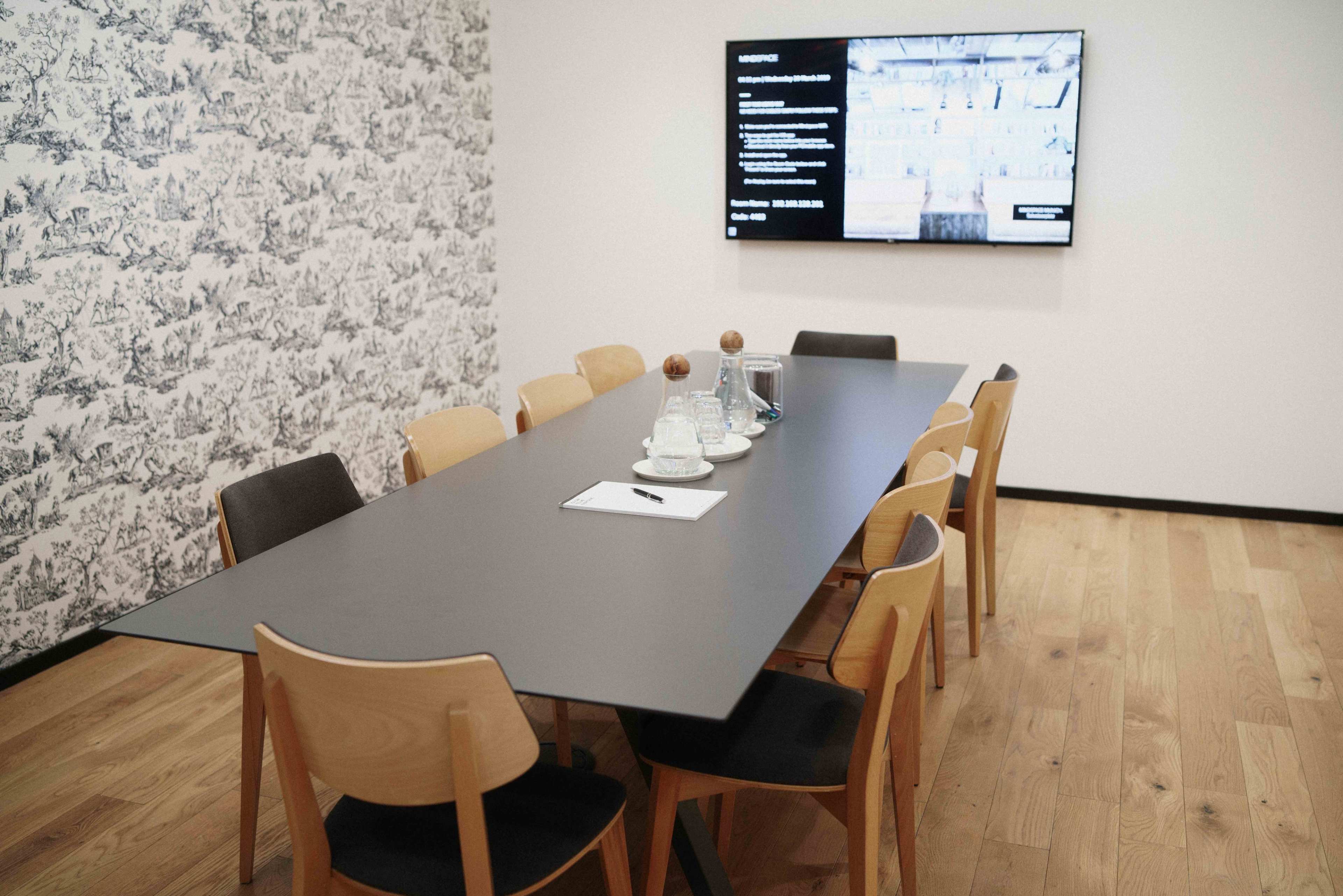 A modern conference room features a long black table surrounded by wooden chairs, with a large screen displaying information on one wall.