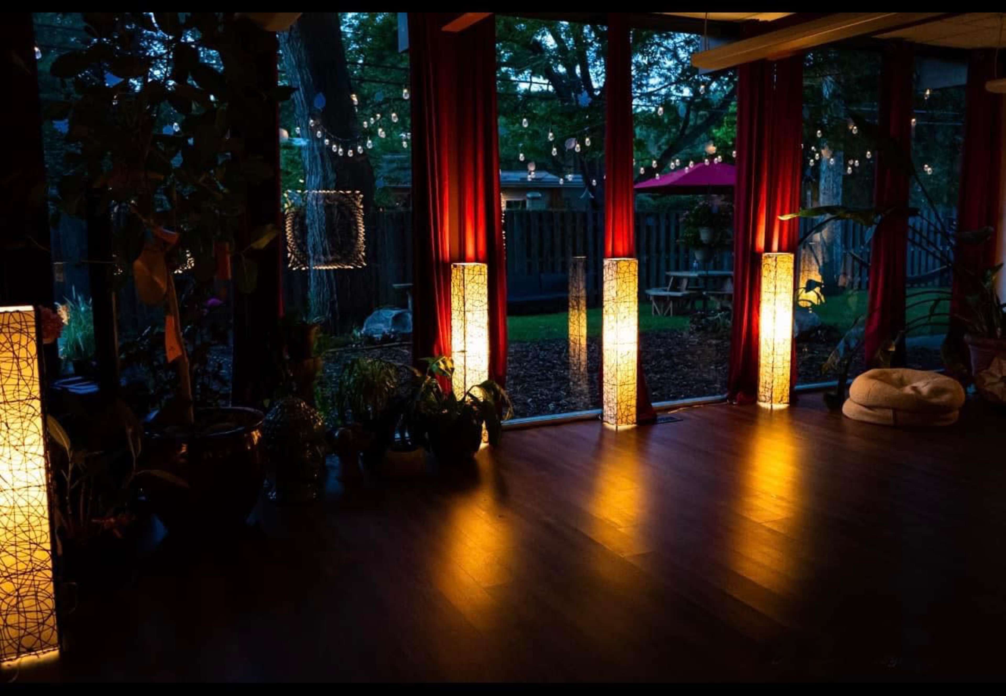 A dimly lit indoor space with decorative light fixtures casting warm illumination, surrounded by plants and large windows revealing an outdoor area.