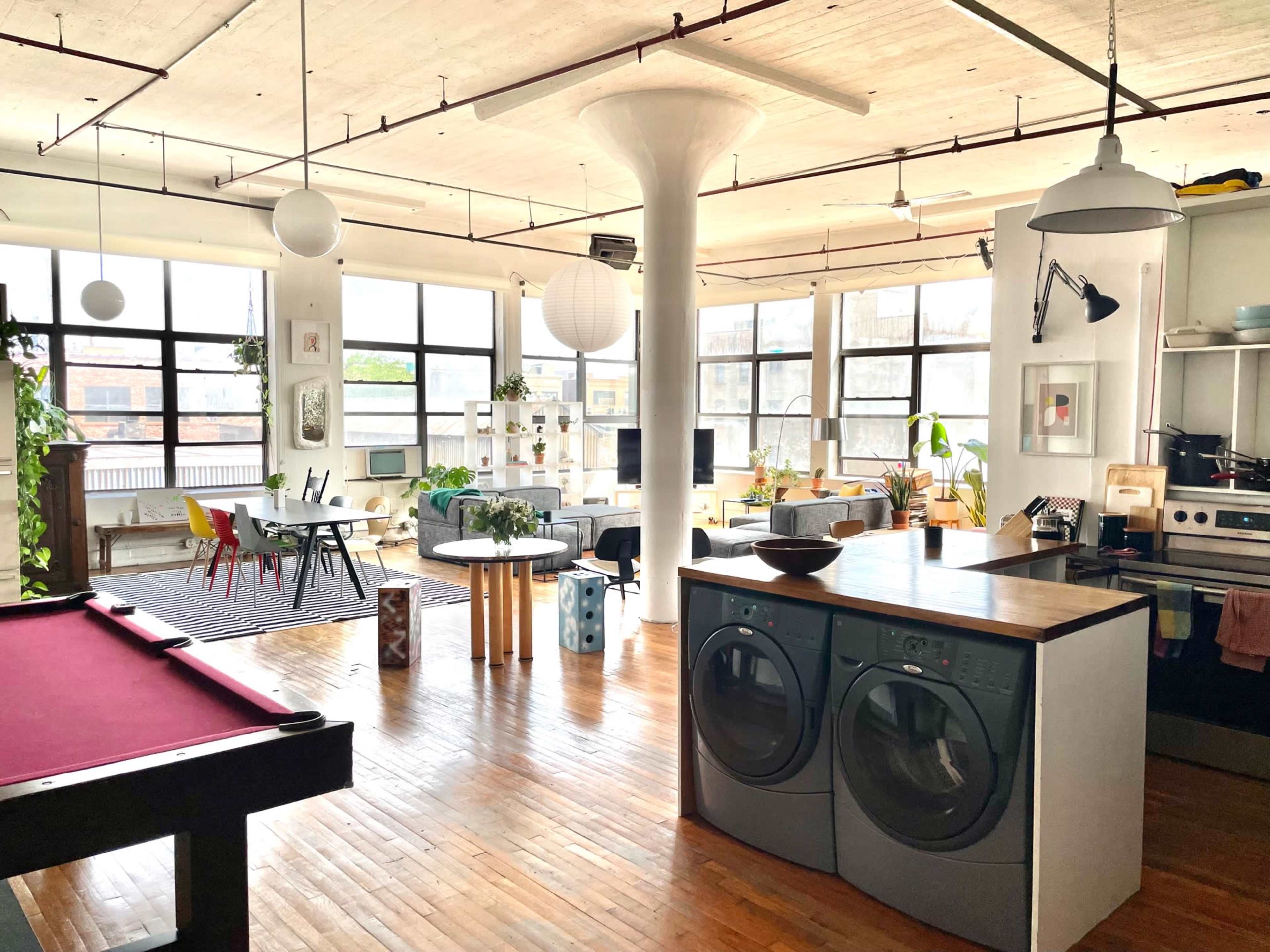 The image shows a spacious, open-concept loft with a kitchen area, a laundry machine, seating arrangements, and large windows that let in natural light.
