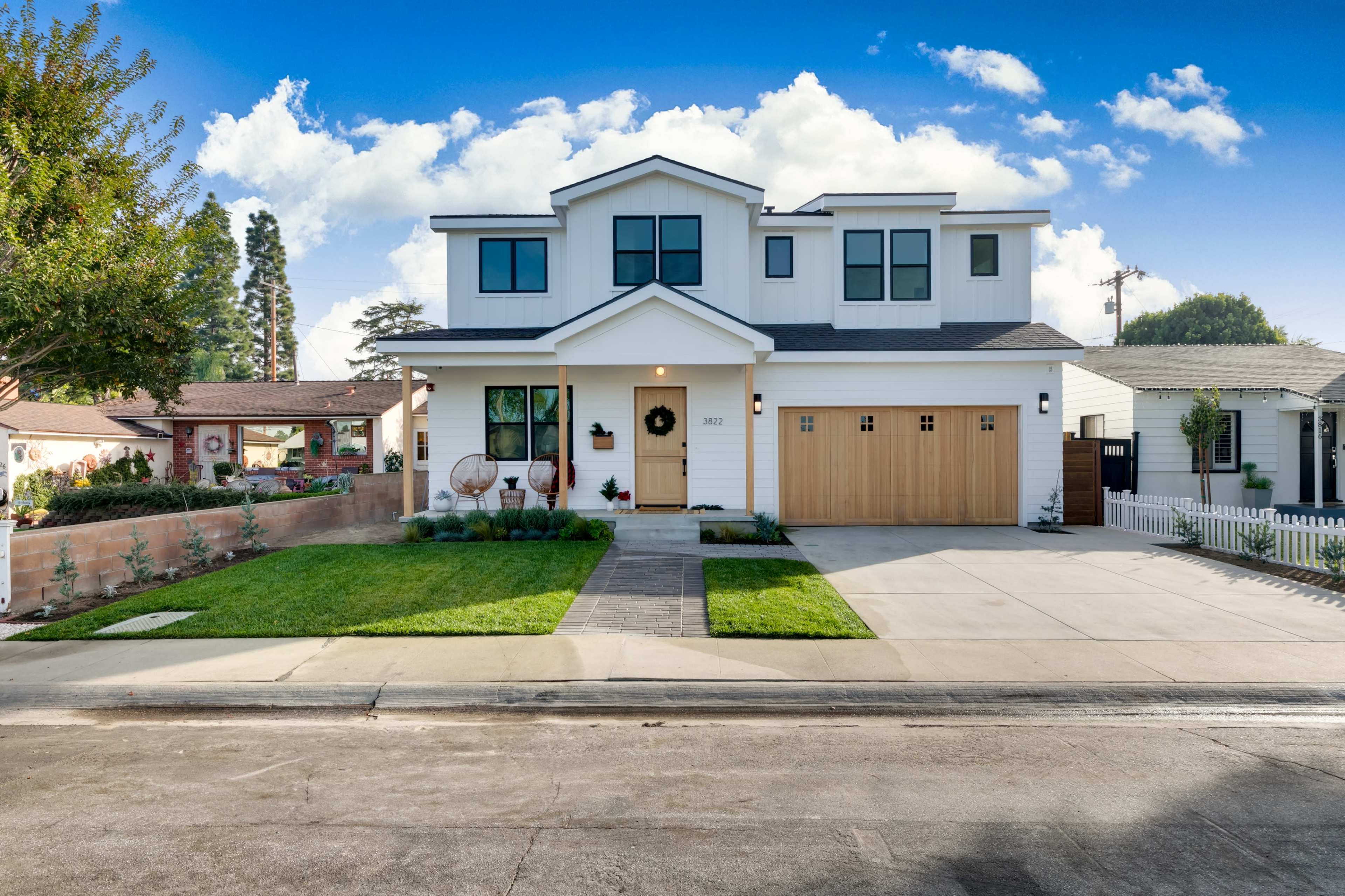 A modern two-story house with a white exterior and a wooden garage door is situated on a residential street, featuring a landscaped front yard and a paved driveway.