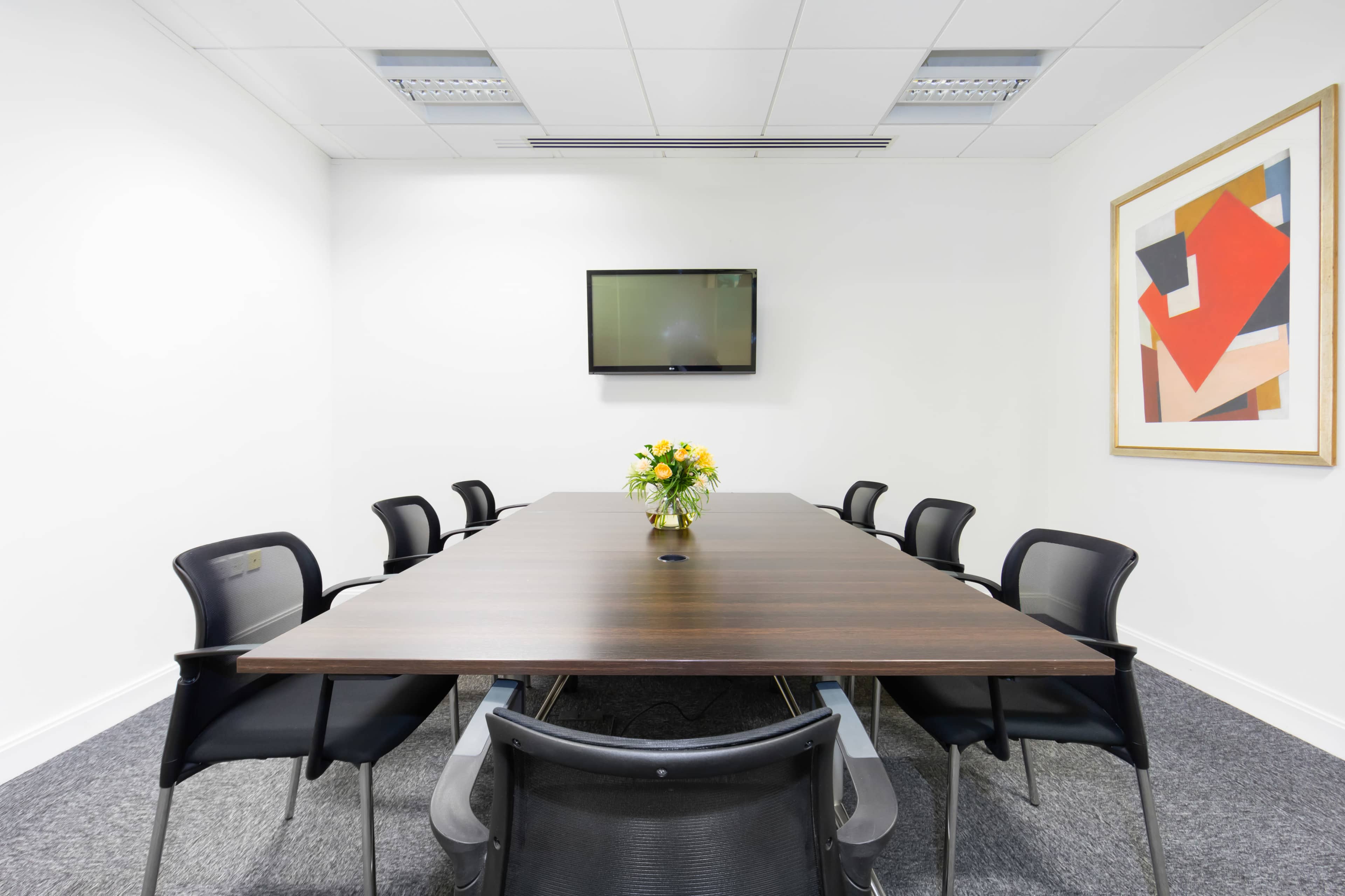A large conference room features a rectangular table with eight chairs, a wall-mounted TV, and a floral centerpiece.