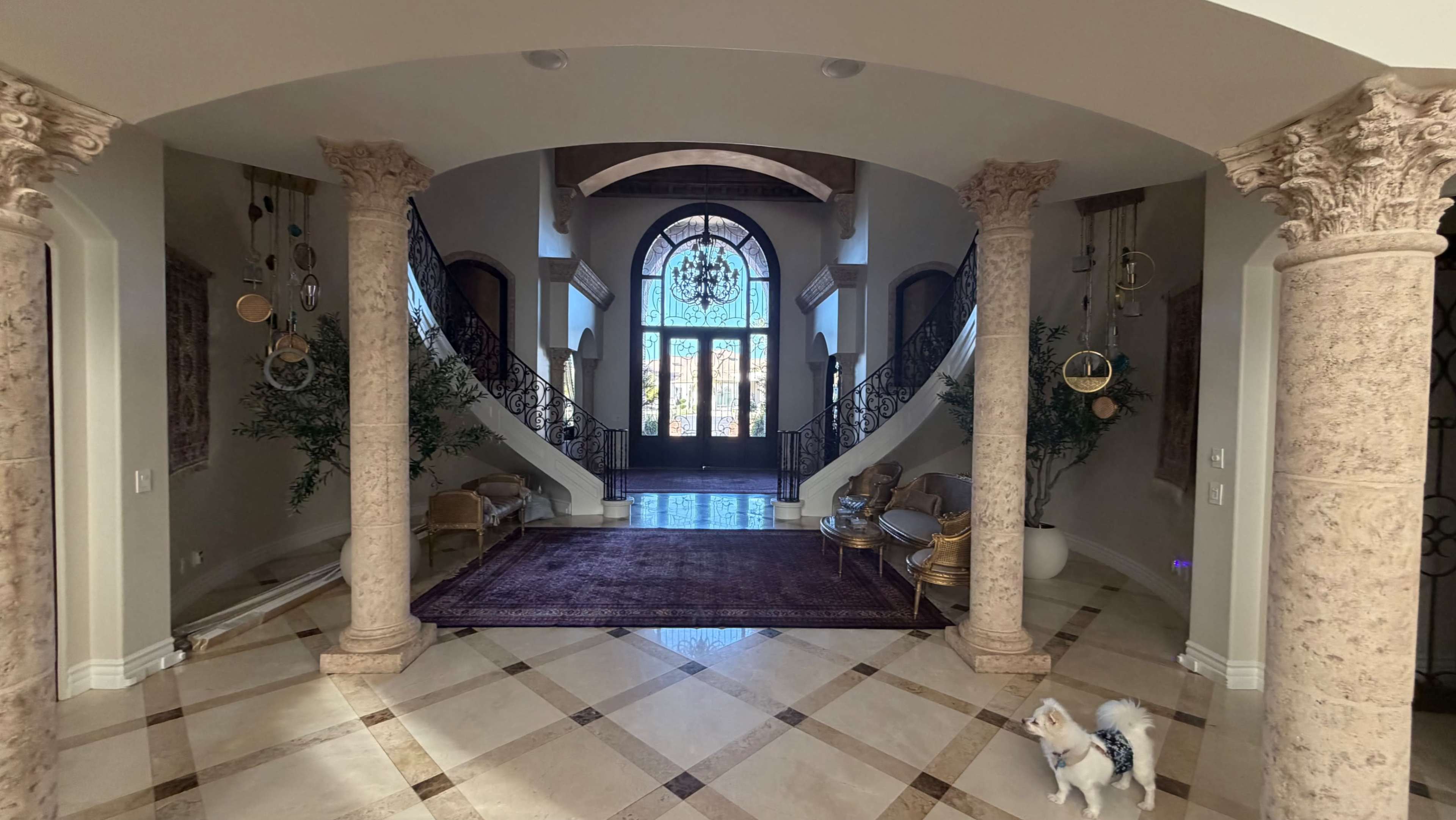The image features a spacious entryway with a grand staircase, ornate columns, and a large arched window providing natural light.