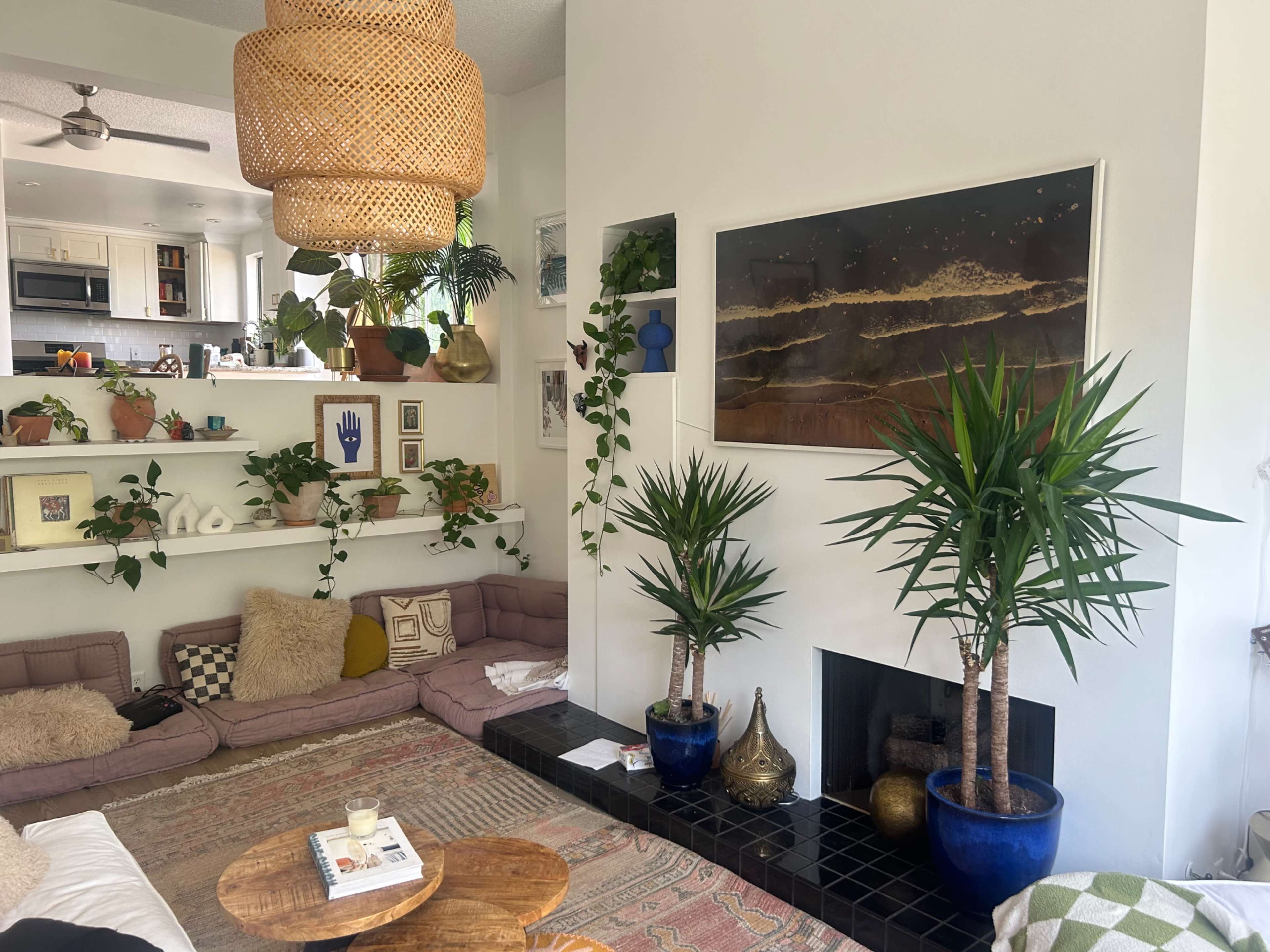 A modern living room with a large wall-mounted painting, two potted plants flanking a fireplace, and a sectional couch arranged around a coffee table.