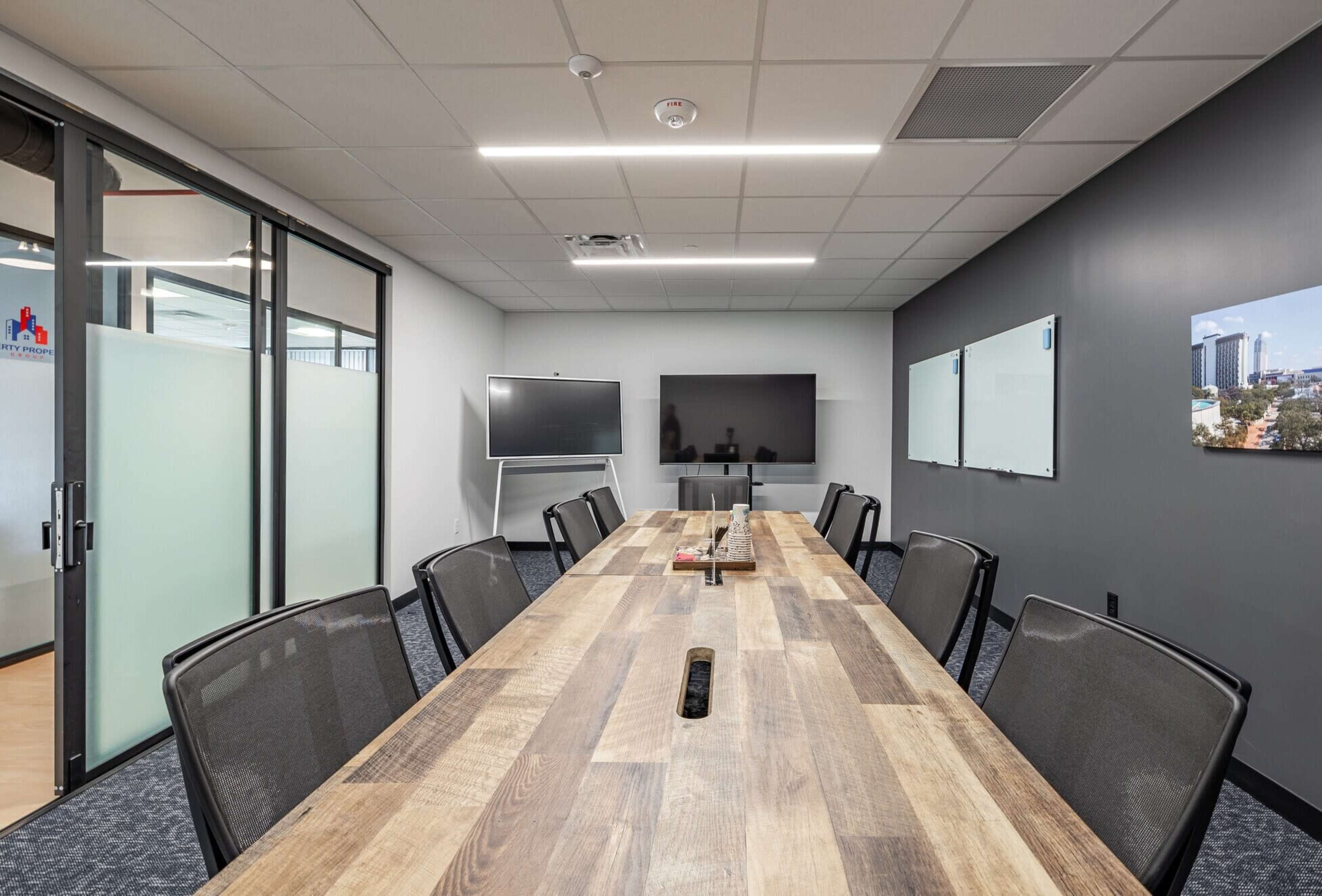 A modern conference room with a long wooden table, black mesh chairs, two large screens, and whiteboards on the walls.