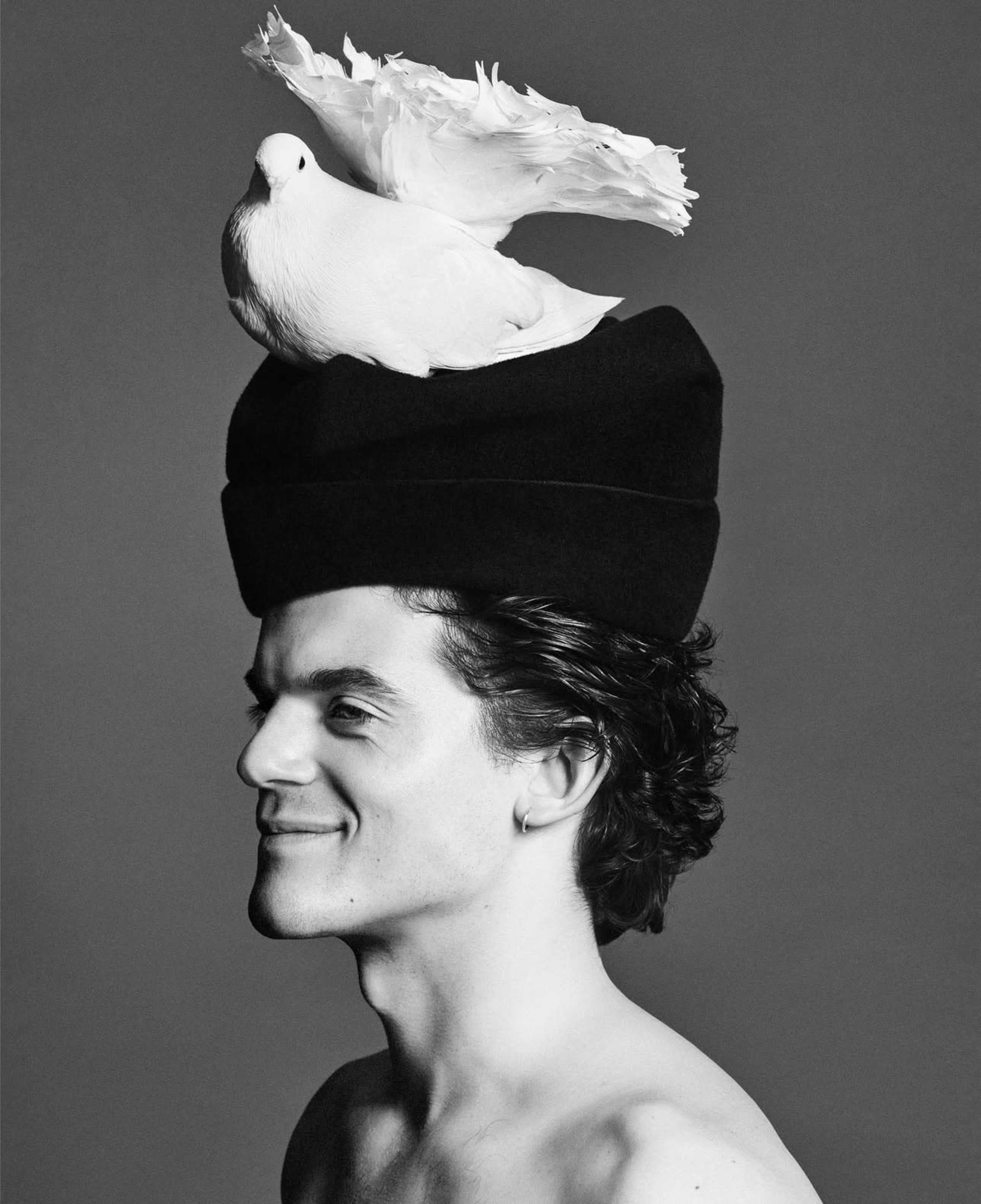 A man with curly hair is posing side profile while wearing a black hat, and a white dove is perched on top of the hat.