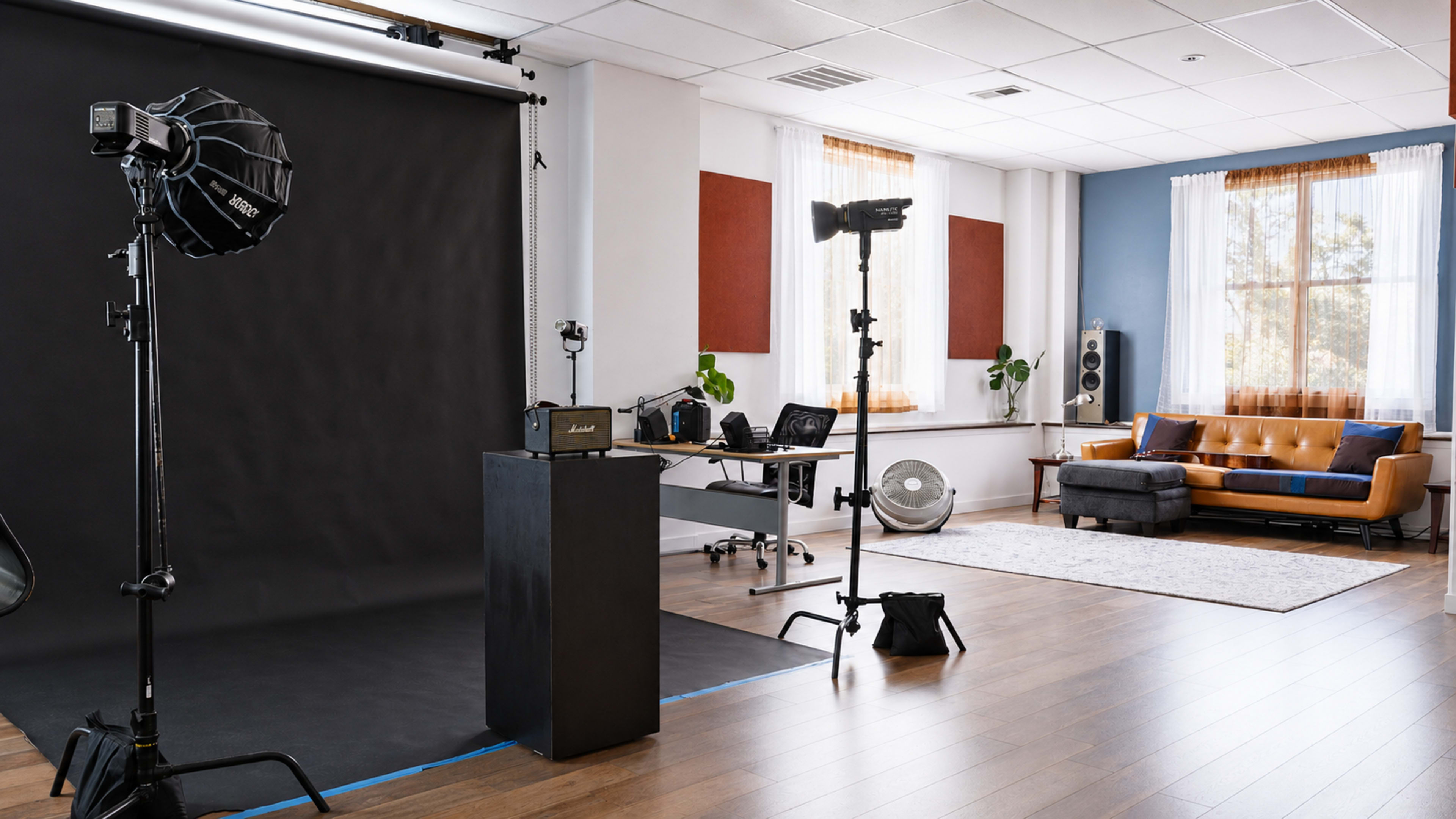 The image shows a photography studio setup featuring a black backdrop, lighting equipment, a desk with a computer, and a couch in a well-lit room with large windows.