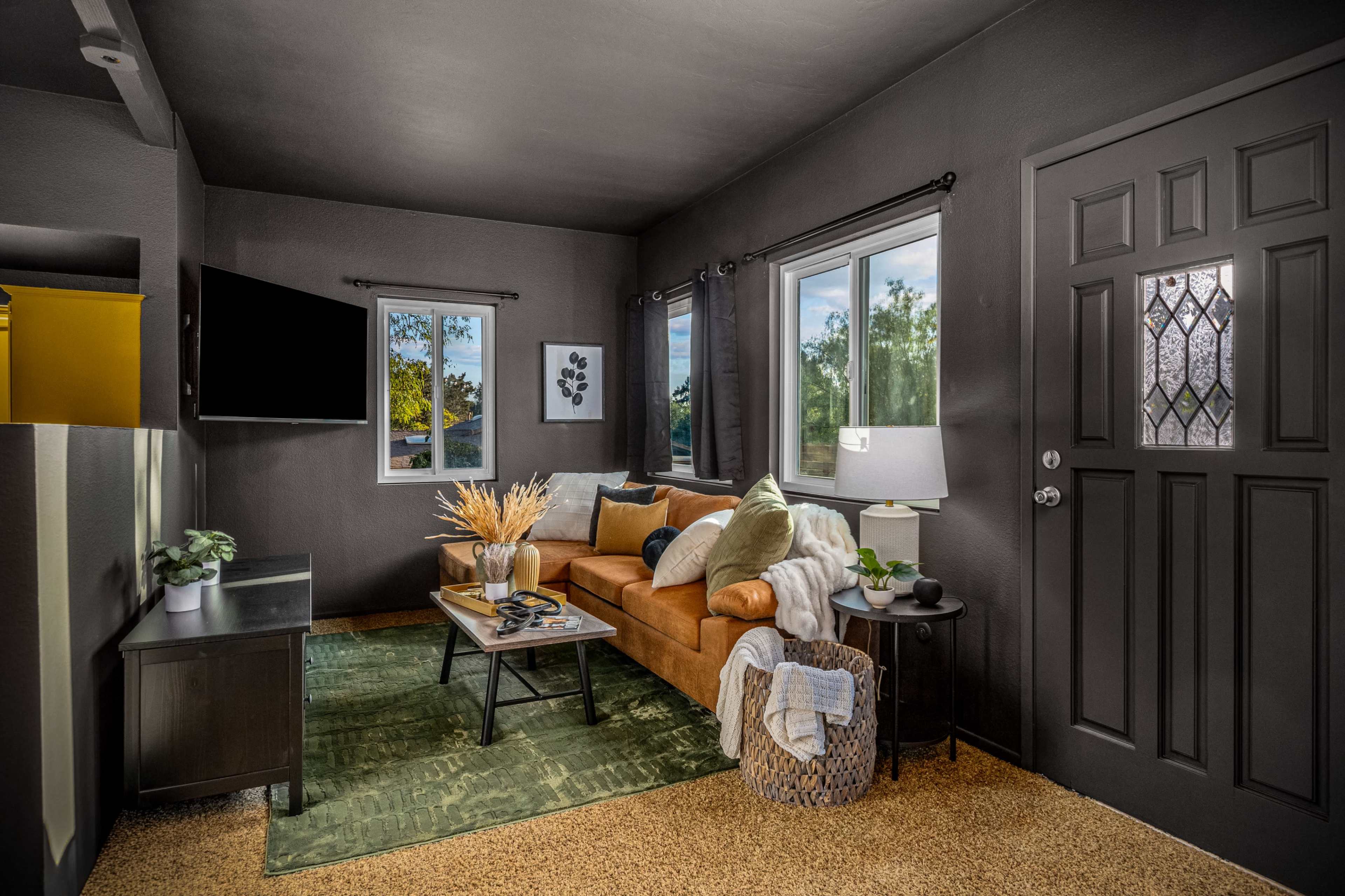 The image shows a living room featuring a brown sofa, a dark accent wall, a window with curtains, a TV mounted on the wall, and a coffee table with decorative items.