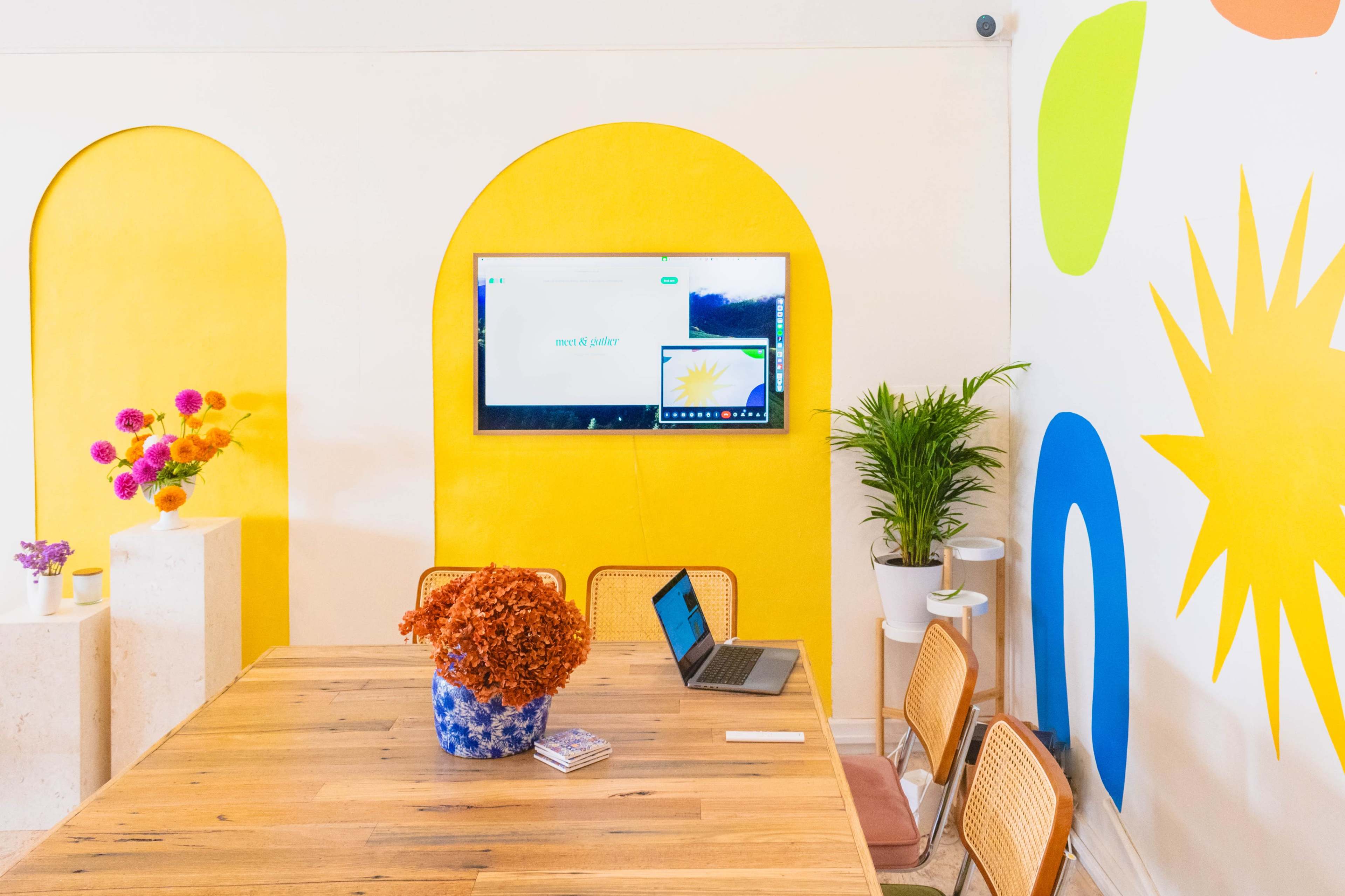 A modern conference room features a wooden table, a laptop, a television displaying a presentation, and brightly colored walls with abstract shapes.