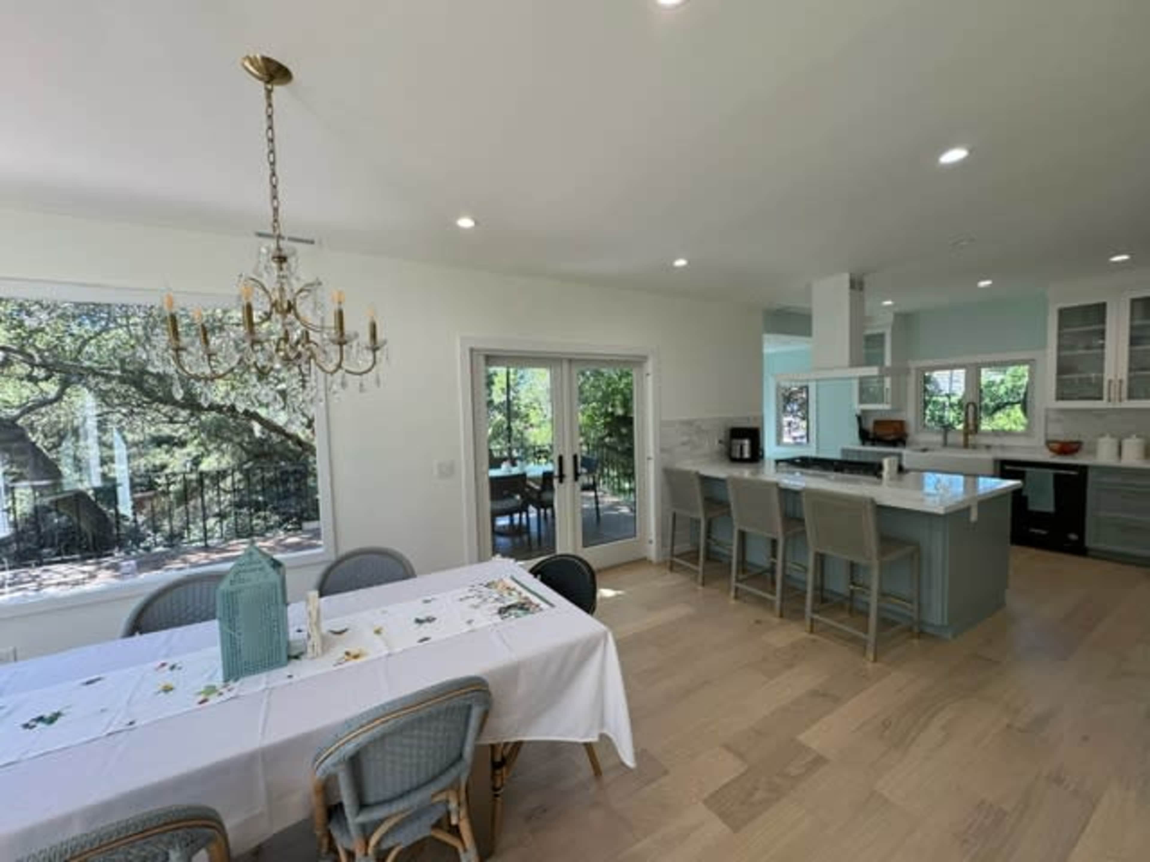 A modern kitchen and dining area with a long dining table and a chandelier, featuring large windows that overlook a tree-filled outdoor space.