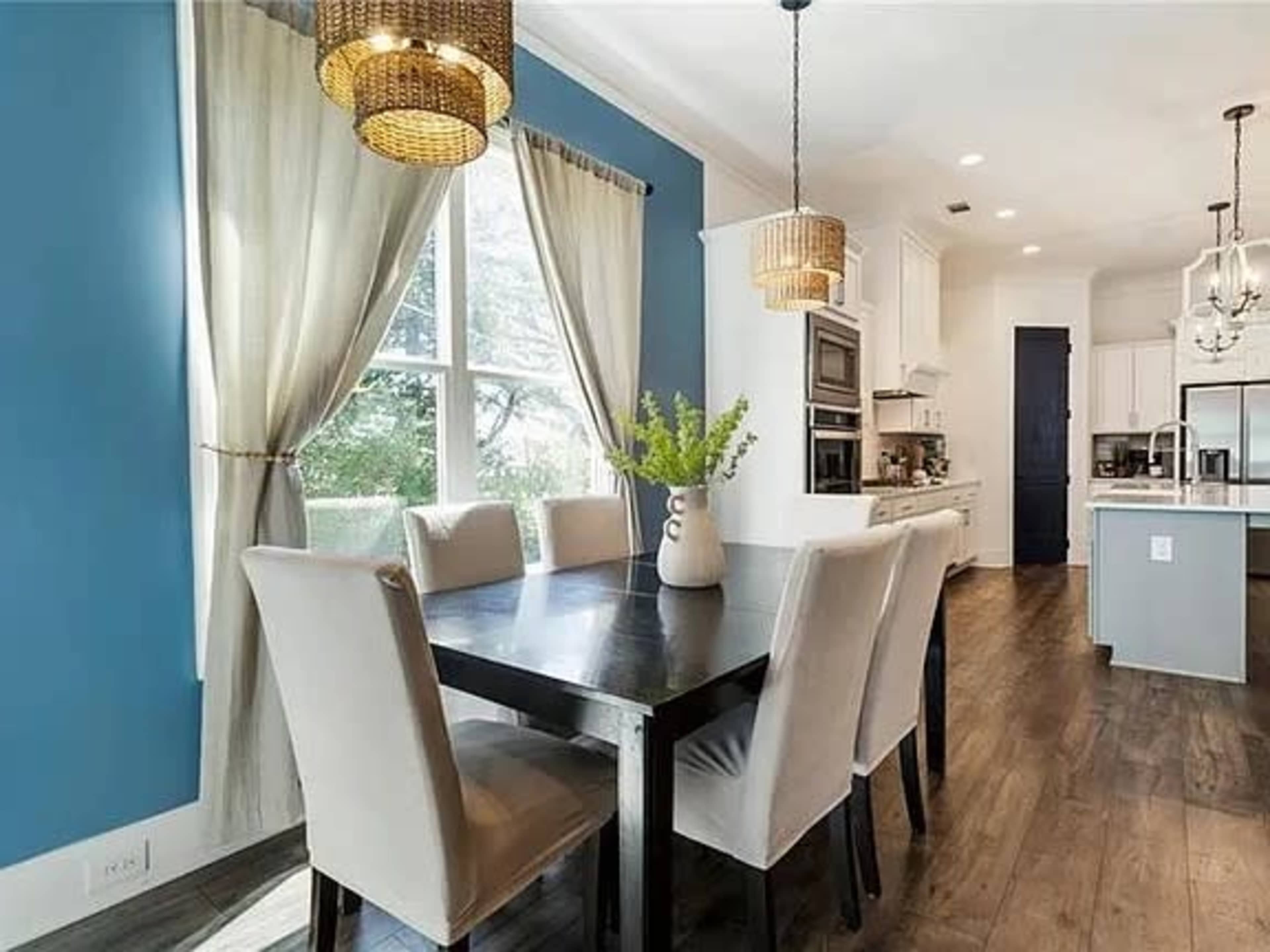 A dining area features a black table surrounded by light-colored chairs, with large windows framed by curtains and modern light fixtures overhead.