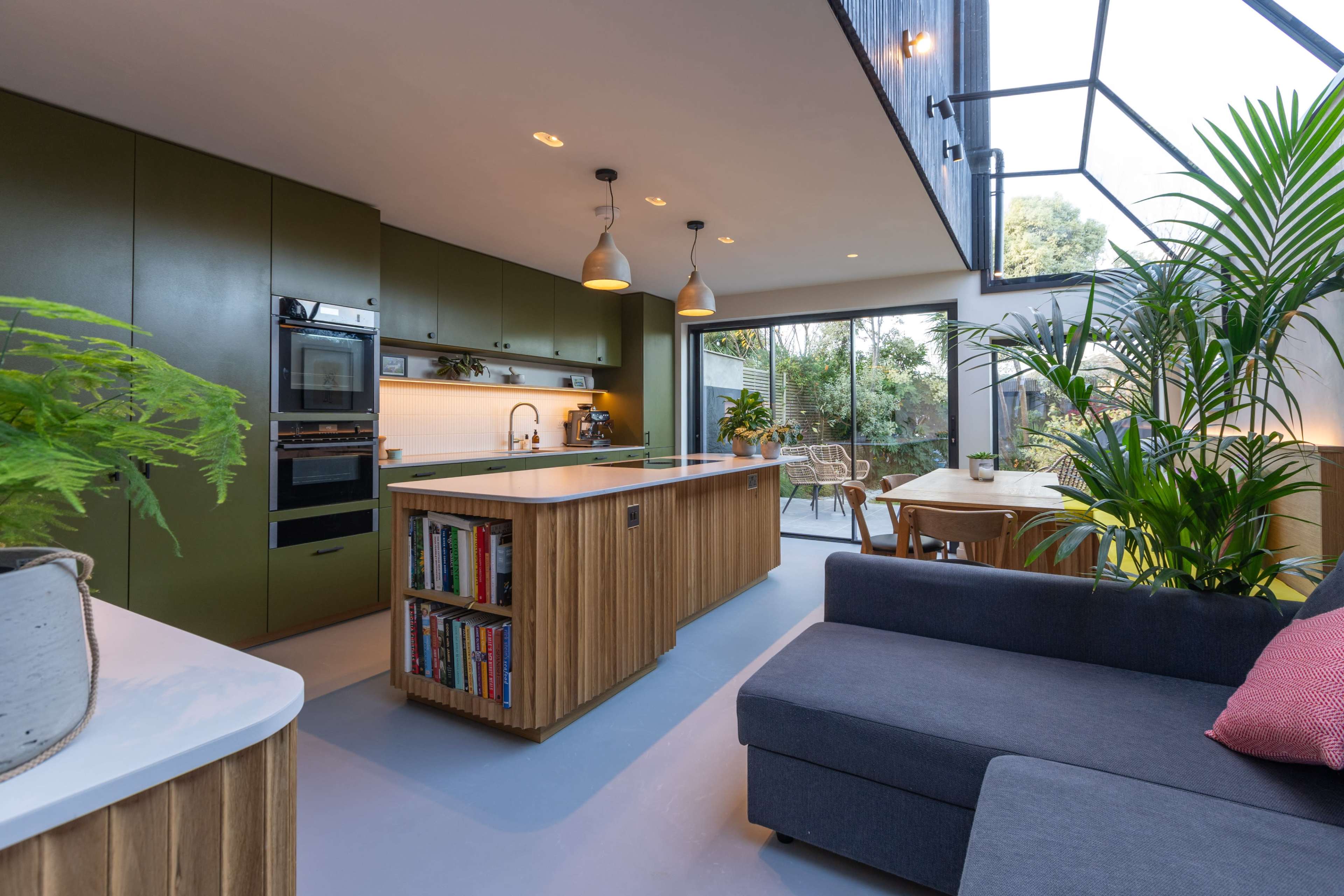 The image shows a modern kitchen with green cabinets, an island with bookshelves, and large windows that open to a garden area.