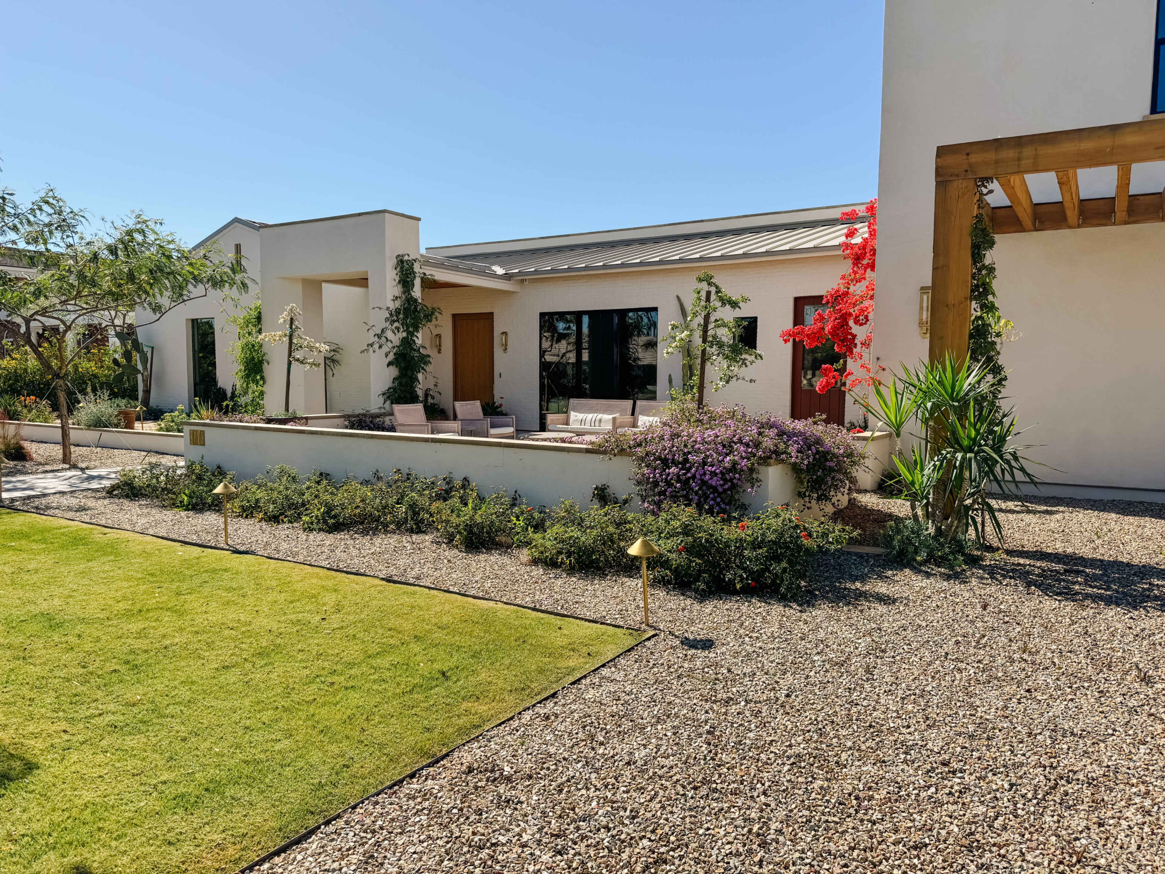 The scene features a modern home with landscaped gardens, including a gravel pathway, green lawn, and colorful flowering plants.