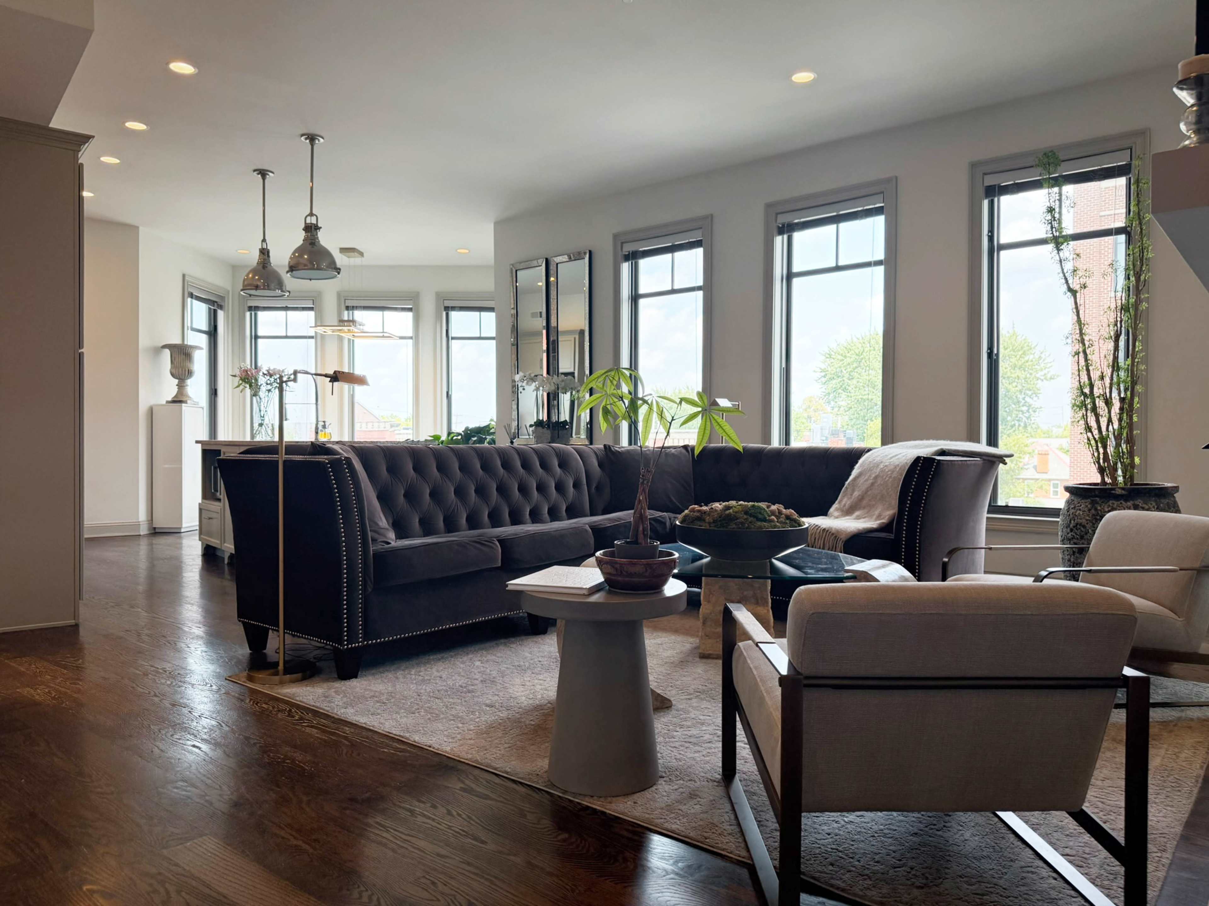 The image shows a modern living room with a dark tufted sofa, a light-colored armchair, a small side table, and large windows allowing natural light to enter.