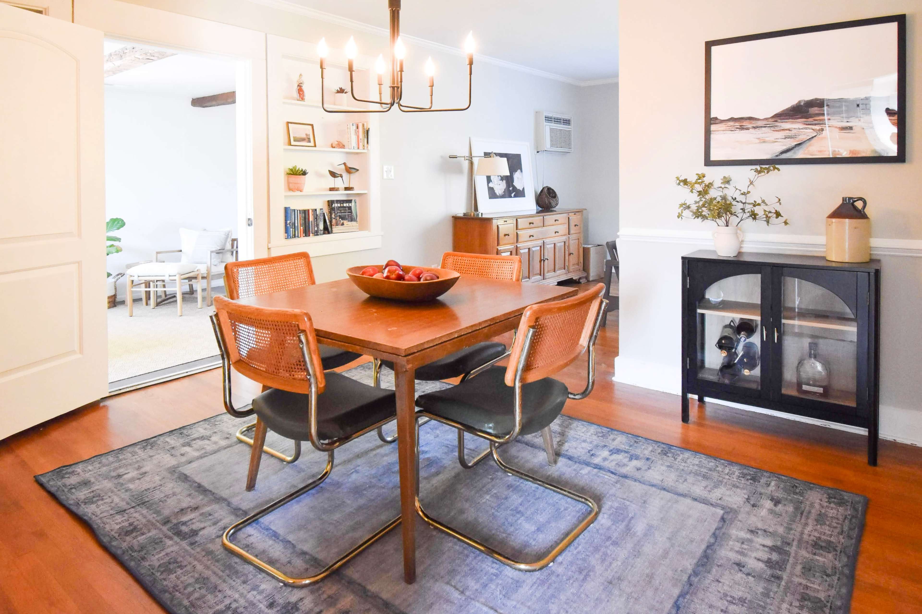 A dining room features a wooden table surrounded by four chairs, with a chandelier above and a bowl of fruit as a centerpiece.