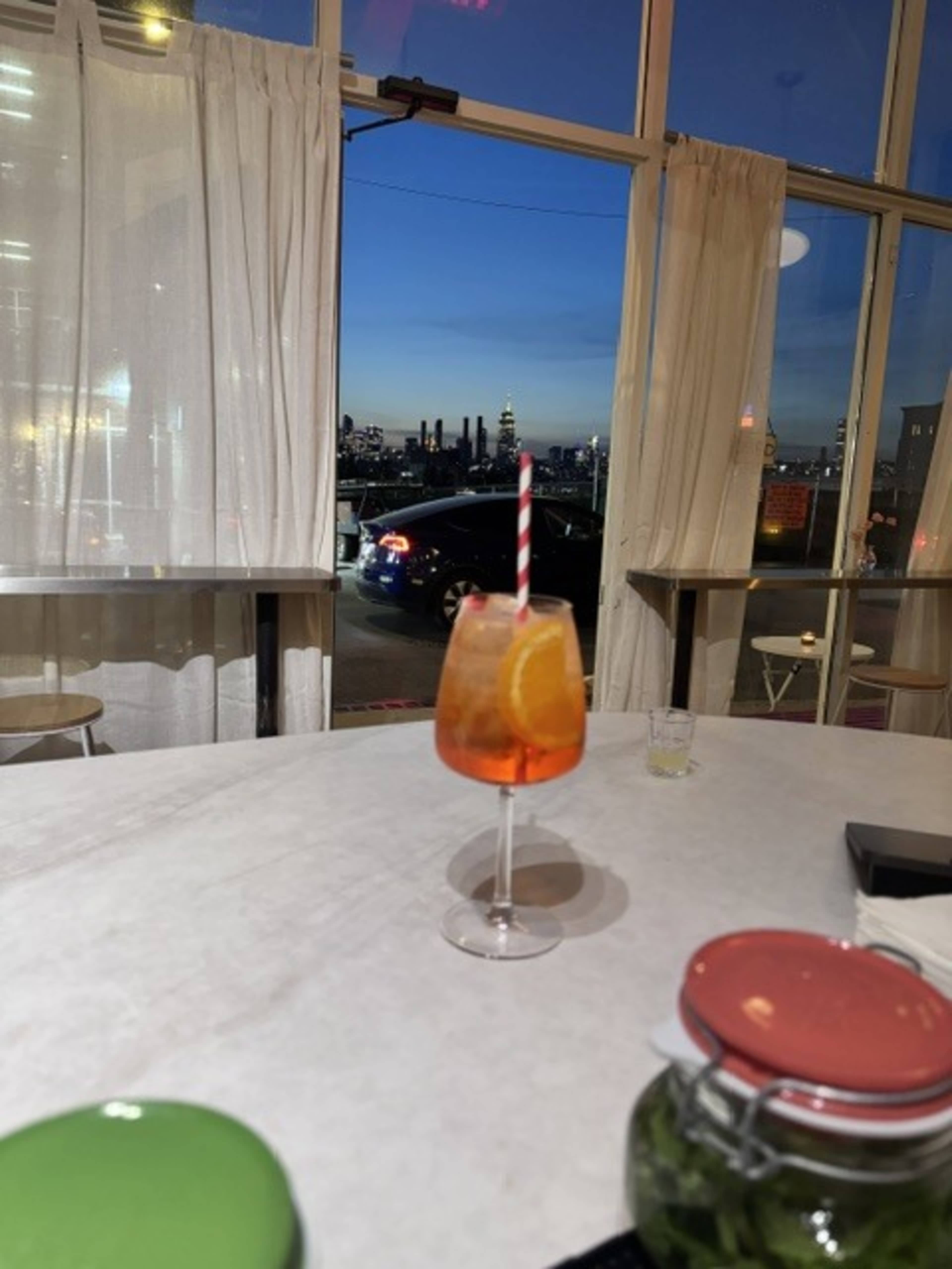 A cocktail glass with a slice of orange and a straw sits on a table, with a city skyline visible through large windows in the background.