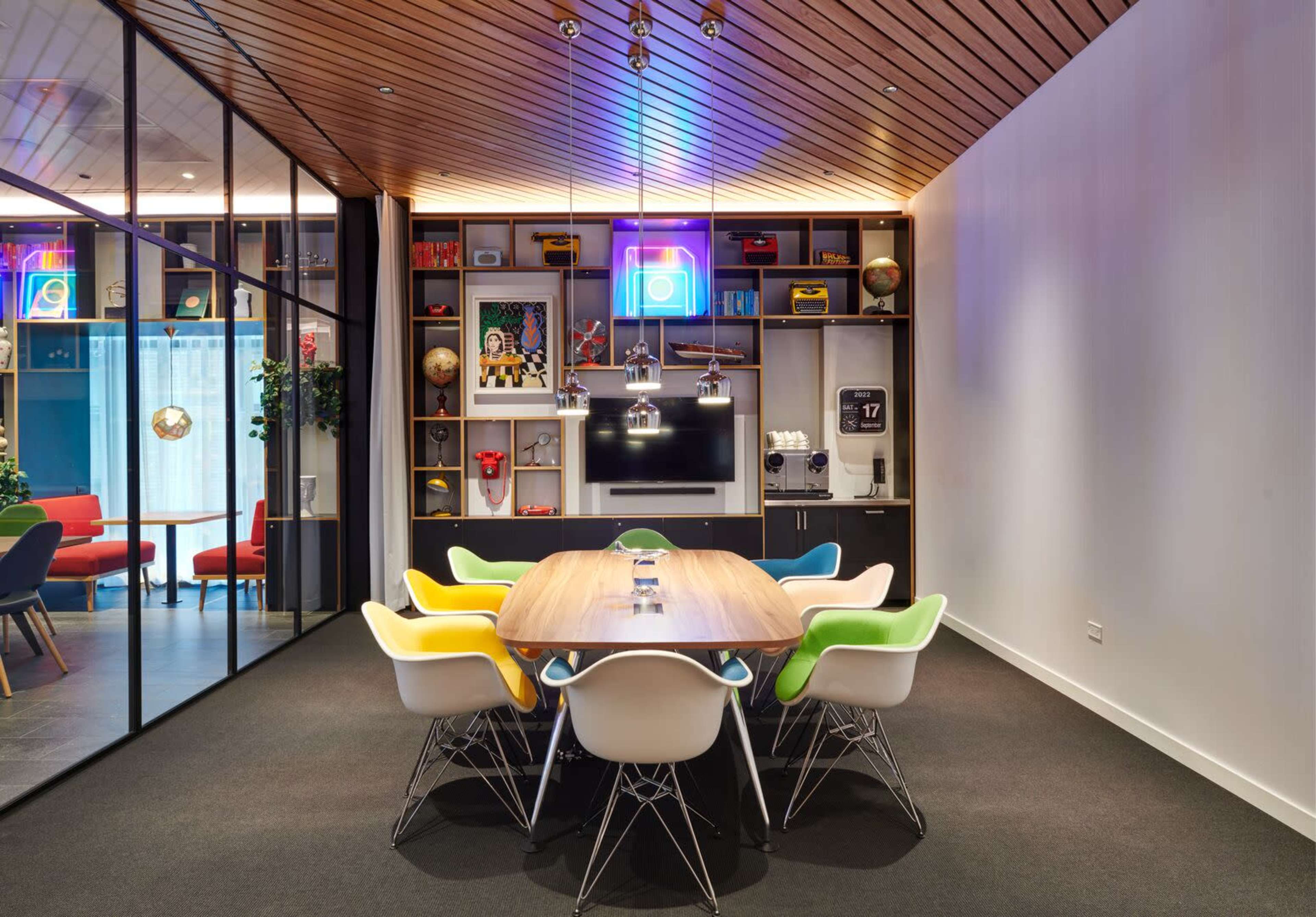 A modern conference room features a large wooden table surrounded by colorful chairs, with shelves displaying various decor items and a television mounted on the wall.