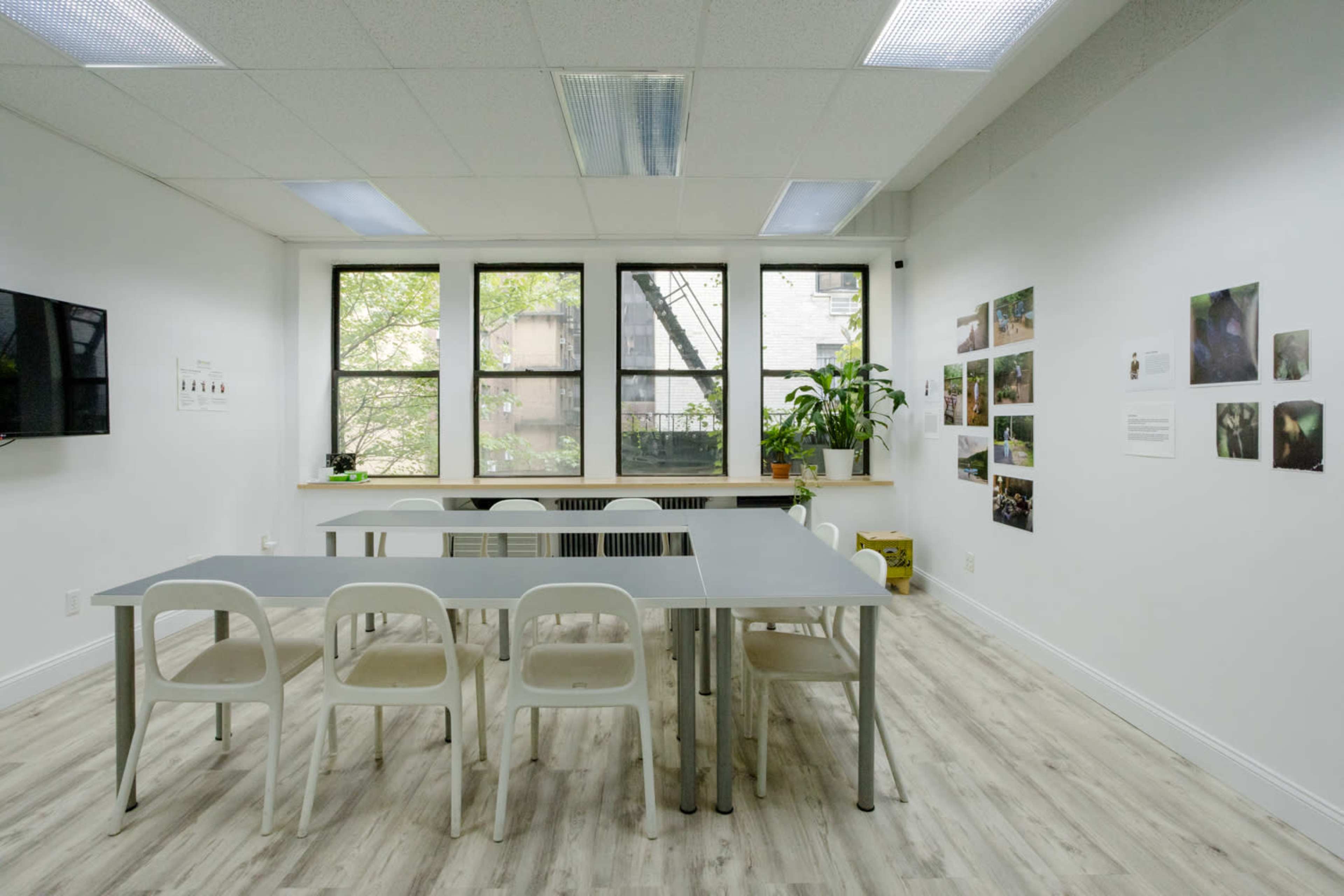 The image shows a modern classroom or meeting space with a long table and white chairs, surrounded by large windows and decorated with plants and wall-mounted photographs.