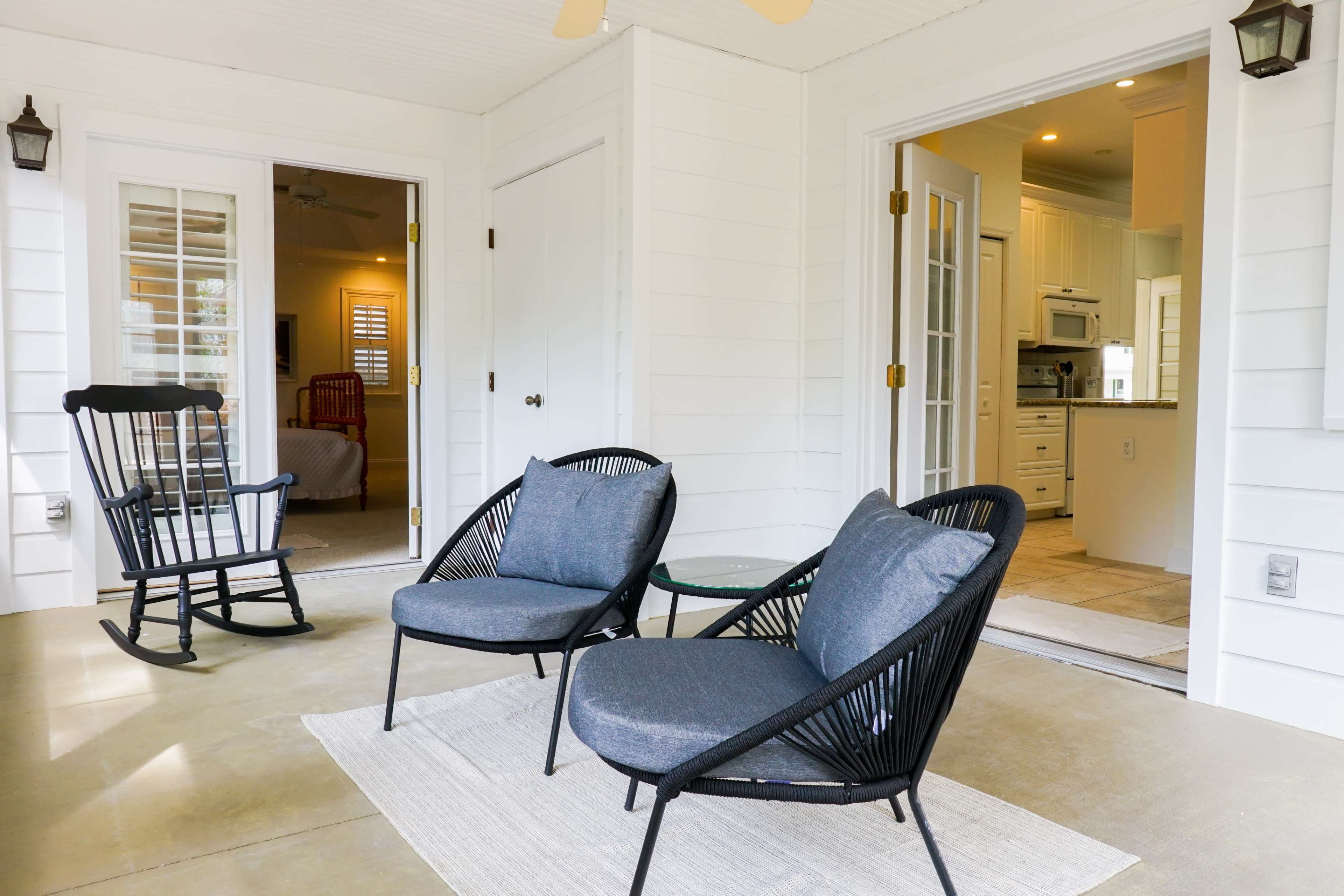 A covered porch with two black lounge chairs, a small glass table, and a rocking chair, all arranged near an entrance to a kitchen.