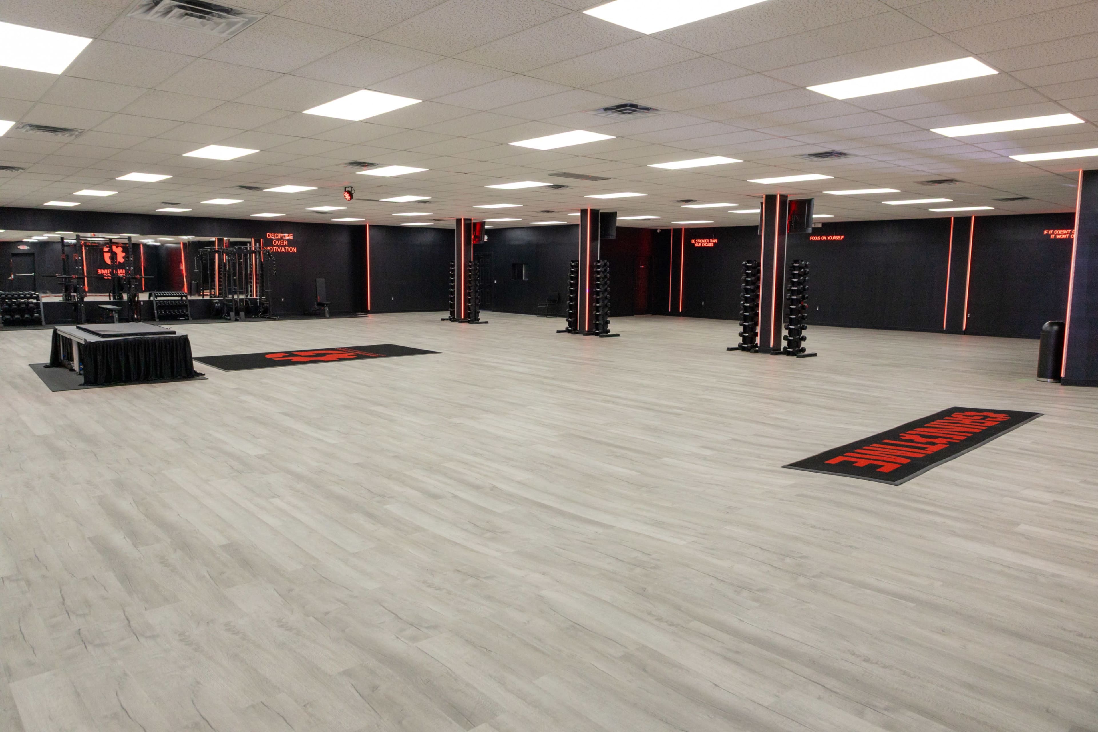 The image shows a spacious gym facility featuring gray flooring, red and black accents, and various workout equipment along the walls.