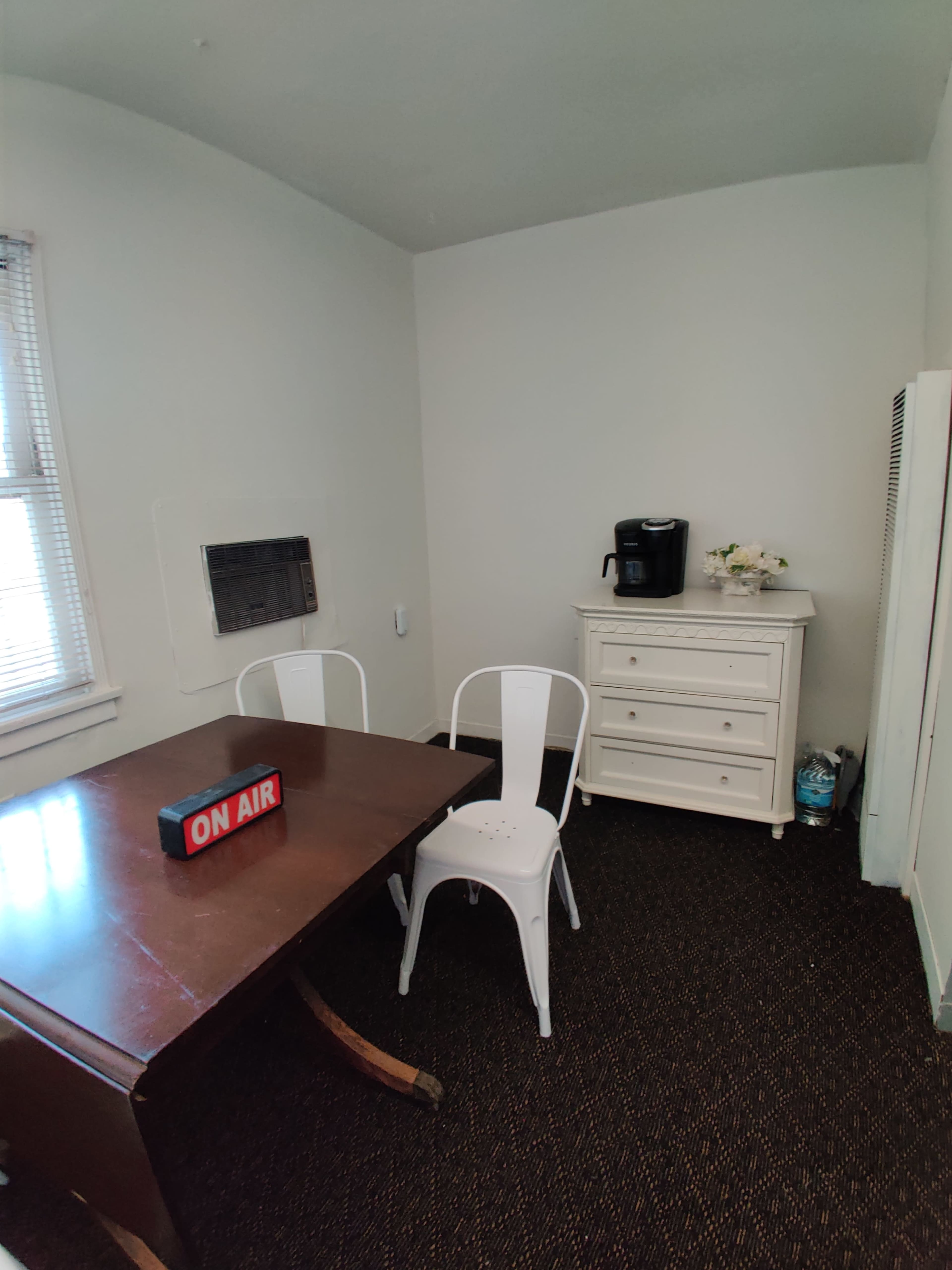 A small room features a wooden table with two white chairs, a coffee maker on a dresser, and a sign reading "ON AIR" on the table.