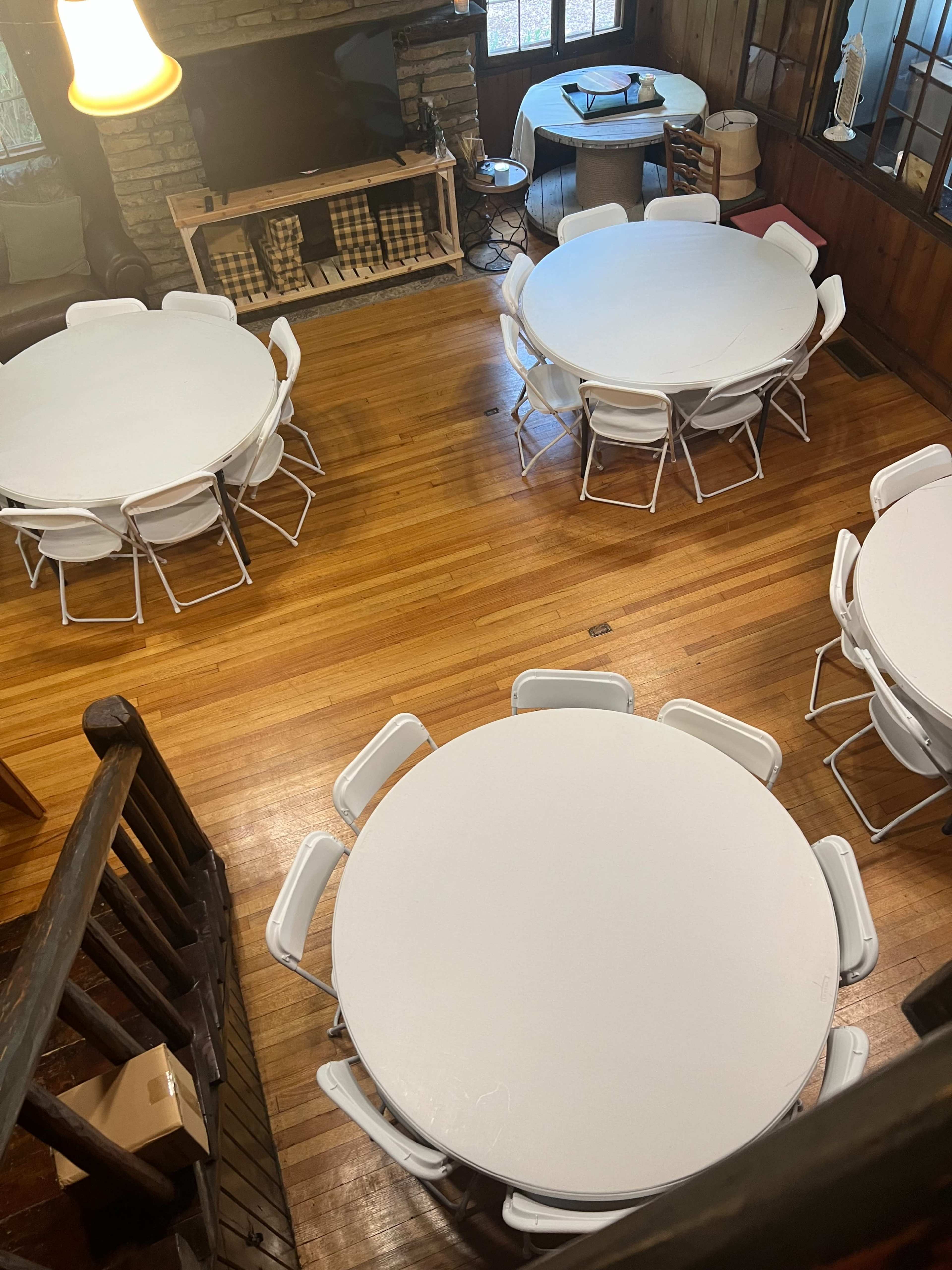 The image shows a spacious room with several round tables and white chairs arranged on a polished wooden floor, along with a stone fireplace in the background.