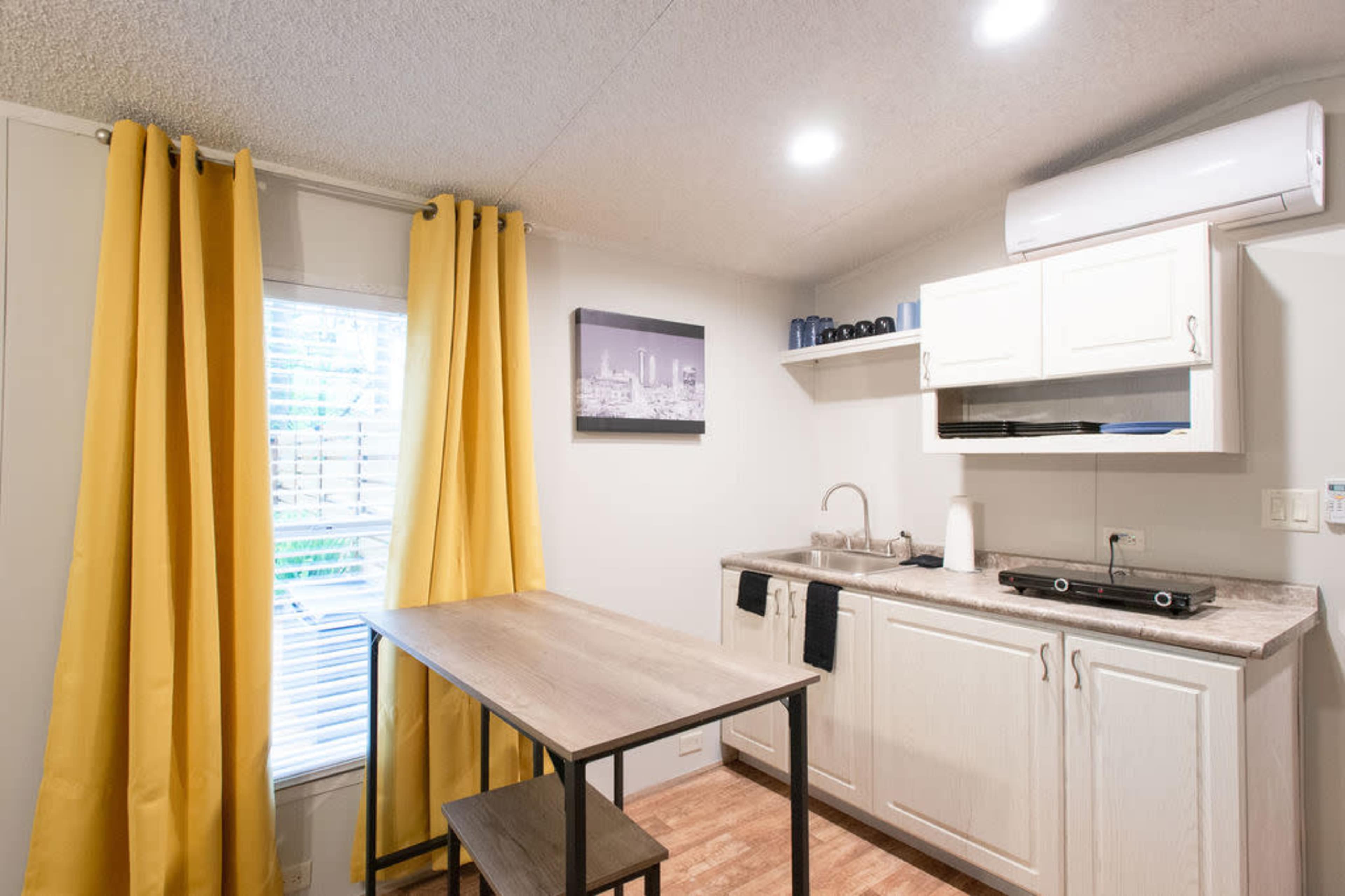 A small kitchen area with a table, two chairs, yellow curtains, and a wall-mounted air conditioning unit.